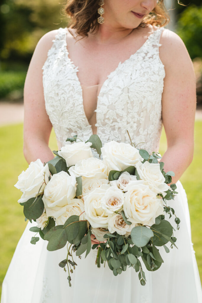 Close-up of a bride holding a lush bouquet of white roses and eucalyptus, showcasing her lace wedding dress with a deep V-neckline at a spring wedding at Daniel Stowe Botanical Garden in Belmont, NC. Captured by DeLong Photography near Charlotte, North Carolina, this botanical garden wedding portrait highlights timeless bridal details and elegant floral design.