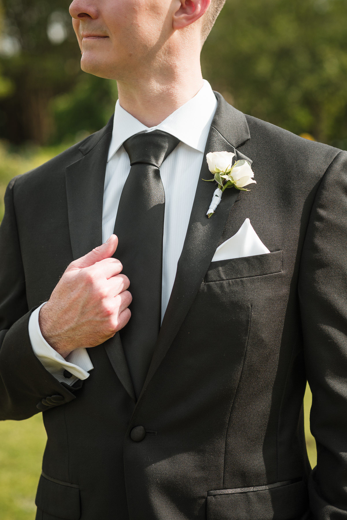 Close-up portrait of groom adjusting his black tie with white rose boutonniere at a spring wedding at Daniel Stowe Botanical Garden in Belmont, NC.