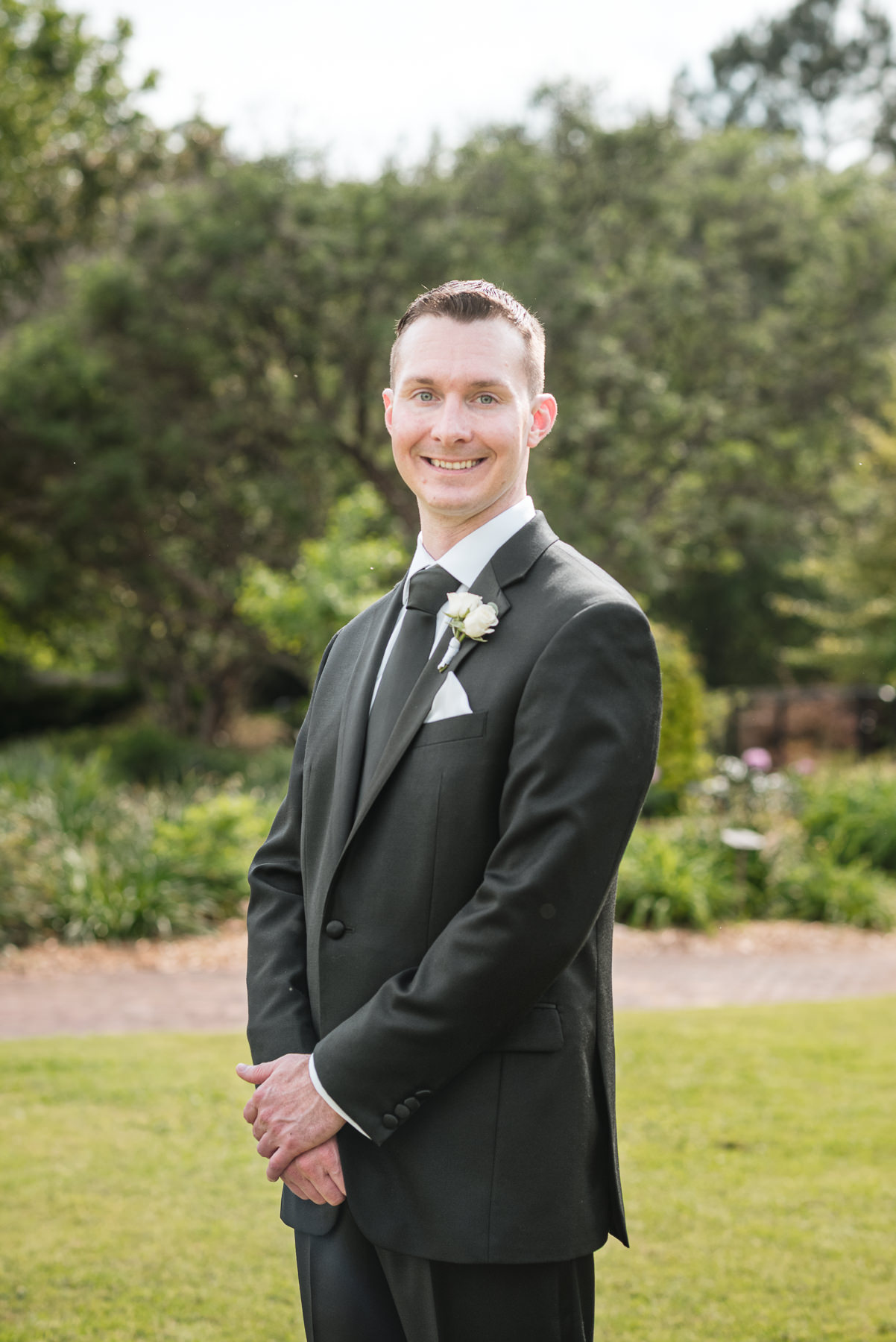 Groom portrait at Daniel Stowe Botanical Garden in Belmont, NC during a spring wedding, wearing a classic black suit with white rose boutonniere and standing on the garden lawn surrounded by lush greenery.