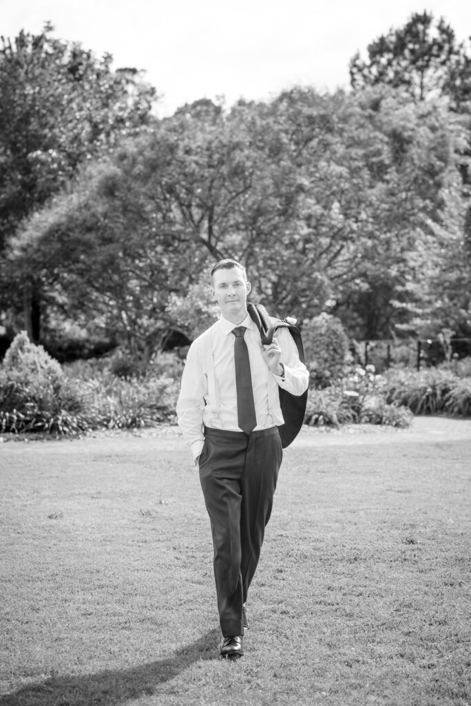 Black and white portrait of a groom walking across the lawn at Daniel Stowe Botanical Garden in Belmont, NC during a spring wedding celebration. This timeless botanical garden wedding image highlights the groom in suspenders and tie with his jacket over his shoulder against a lush garden backdrop.