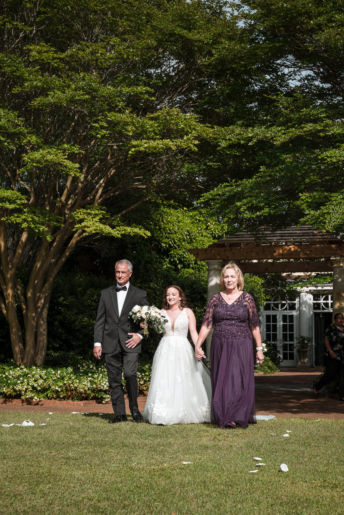 Bride walking down the aisle with her parents during a spring wedding ceremony at Daniel Stowe Botanical Garden in Belmont, NC, surrounded by lush greenery and garden architecture.