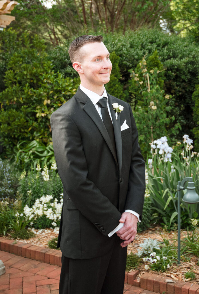 Groom waiting at the outdoor ceremony at Daniel Stowe Botanical Garden in Belmont, NC during a spring wedding, wearing a black suit with white rose boutonniere and standing along a brick pathway surrounded by blooming garden flowers and lush greenery.