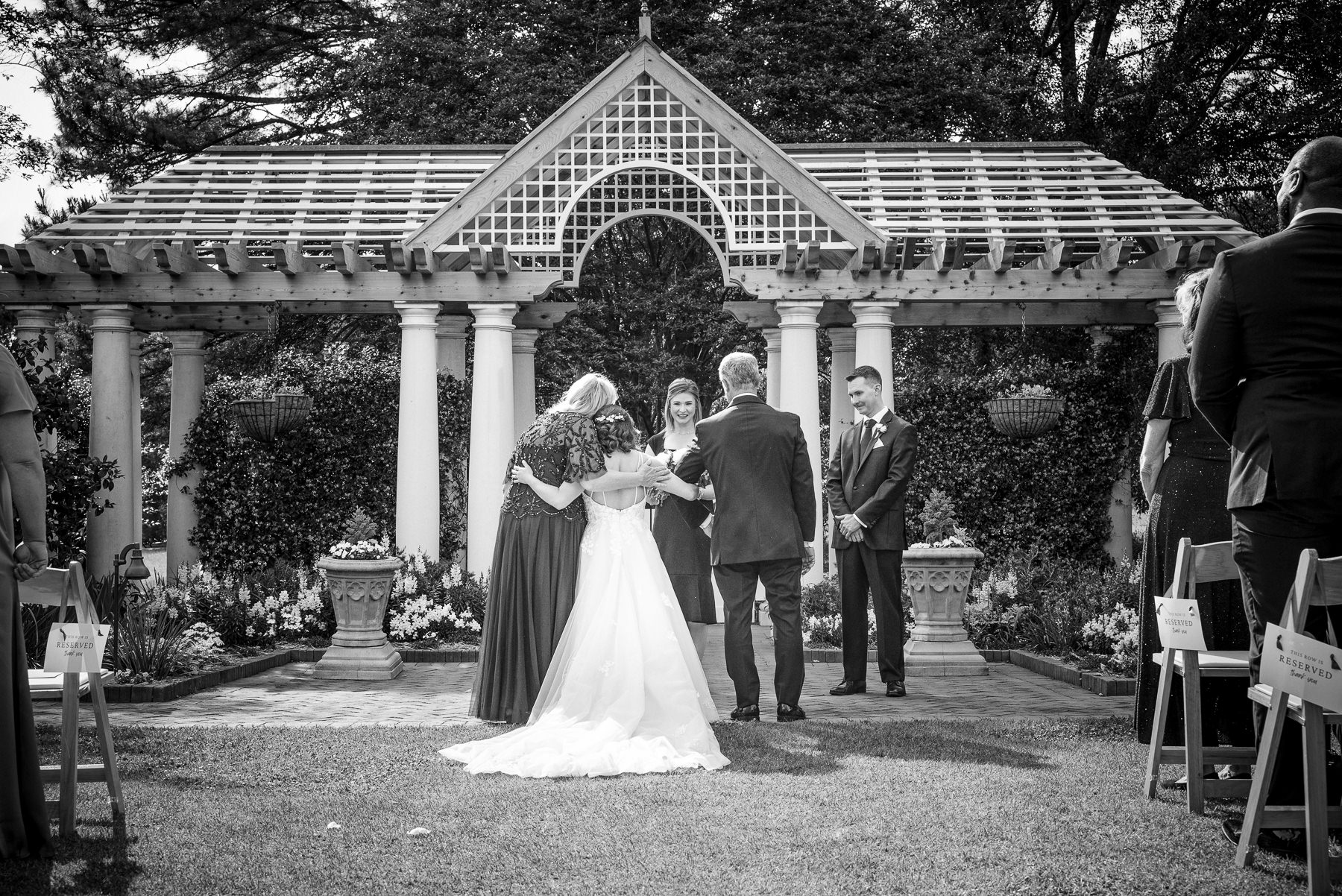 Black and white photo of a spring wedding ceremony at Daniel Stowe Botanical Garden in Belmont, NC, with the bride and groom standing beneath the garden arbor at the Levine Pavilion surrounded by family and blooming flowers.