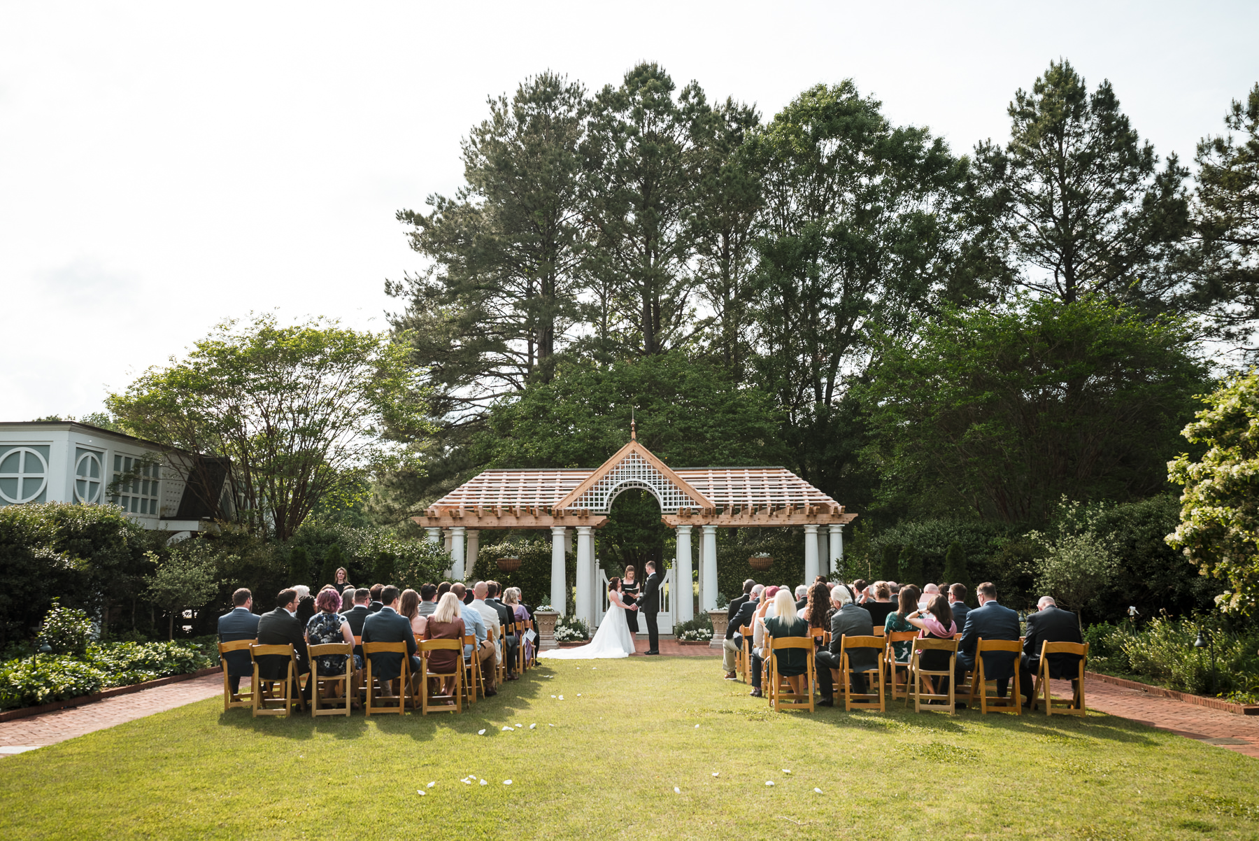 Wide view of a spring outdoor wedding ceremony at Daniel Stowe Botanical Garden in Belmont, NC, with guests seated on the lawn facing the Levine Pavilion arbor surrounded by lush gardens and tall trees.