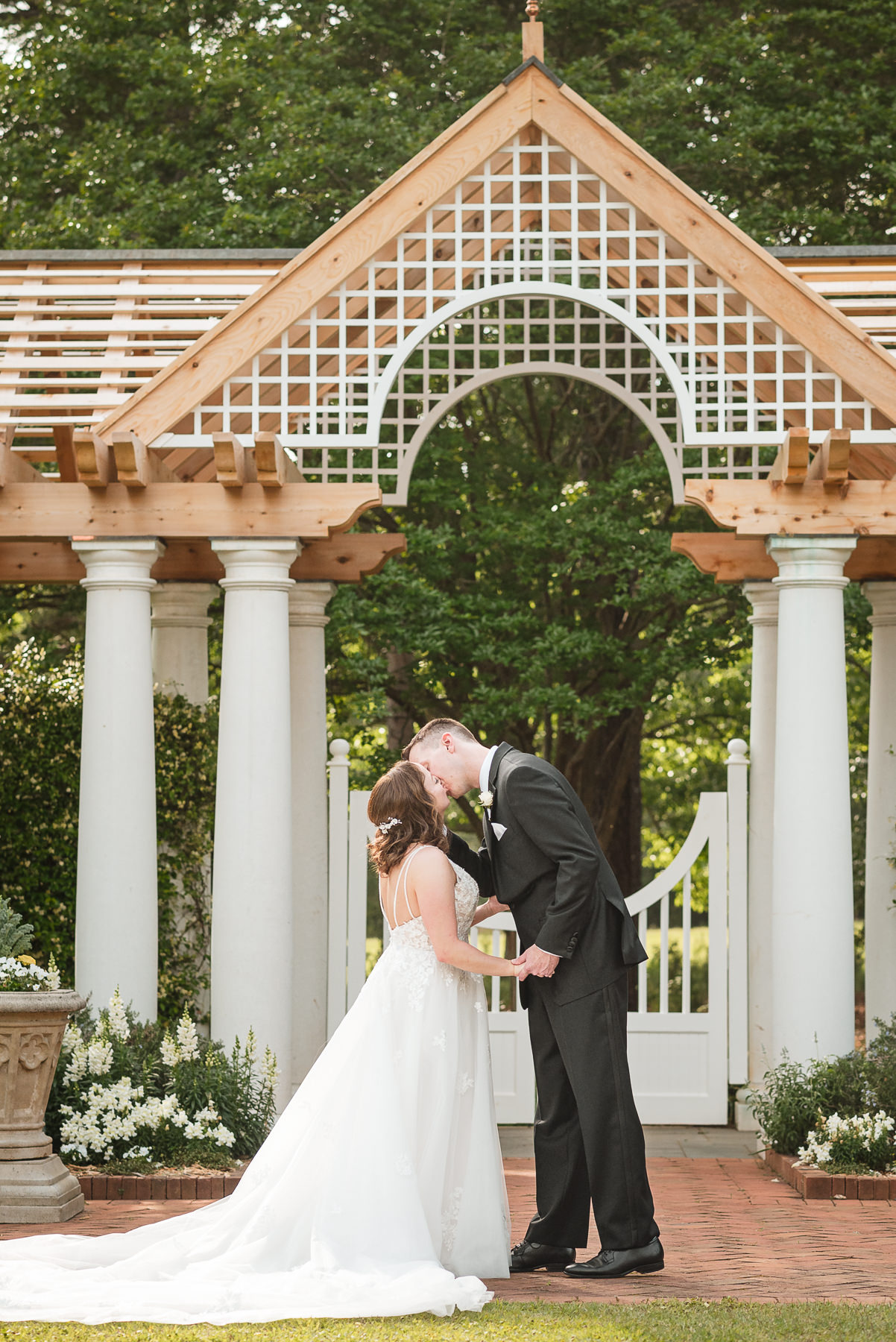 Bride and groom share a romantic kiss beneath the white garden arbor during their spring wedding at Daniel Stowe Botanical Garden in Belmont, NC. Captured by DeLong Photography, this elegant Charlotte-area outdoor wedding features a lace bridal gown, classic black tuxedo, and lush botanical garden backdrop.
