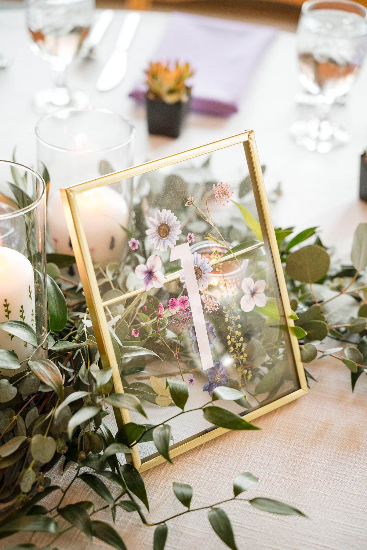 Elegant spring wedding reception table centerpiece featuring a gold-framed pressed floral table number surrounded by greenery and candles at Daniel Stowe Botanical Garden in Belmont, NC. Captured by DeLong Photography, this Charlotte-area garden wedding showcases romantic botanical decor and refined outdoor reception details.