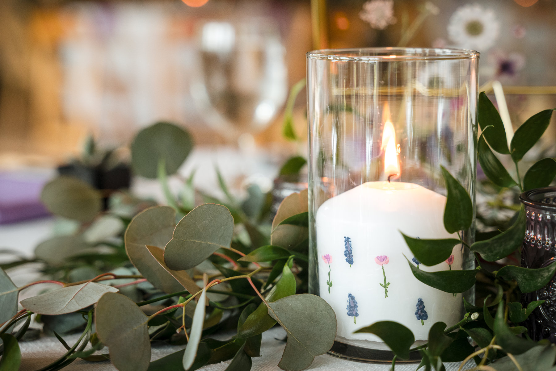 Close-up of a custom wedding candle with hand-painted flowers inside a glass hurricane vase, surrounded by eucalyptus greenery at a spring wedding reception at Daniel Stowe Botanical Garden in Belmont, NC.