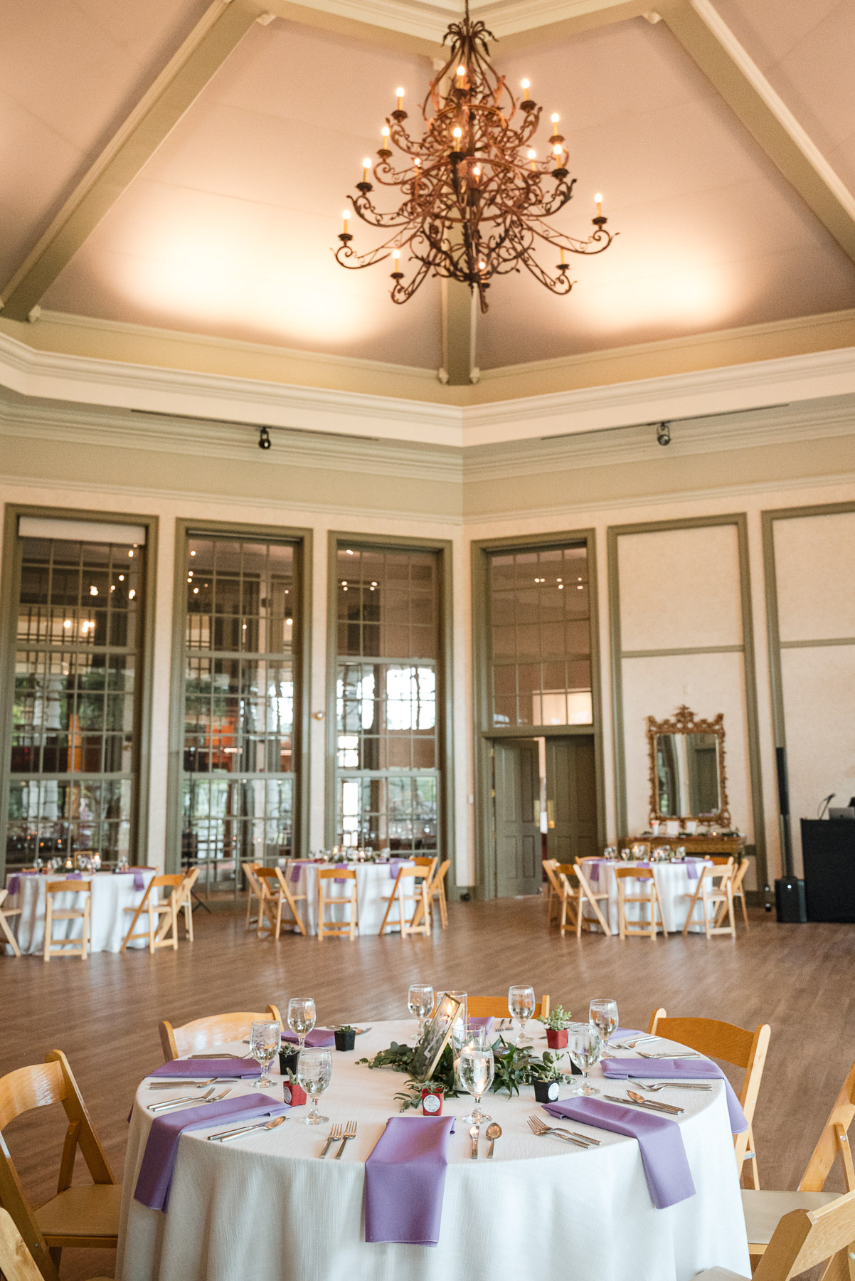 Elegant spring wedding reception setup inside Daniel Stowe Botanical Garden in Belmont, NC, featuring round tables with purple linens, greenery centerpieces, and a grand chandelier overhead. Captured by DeLong Photography, this Charlotte-area garden wedding venue showcases classic architecture, tall windows, and refined indoor reception decor.