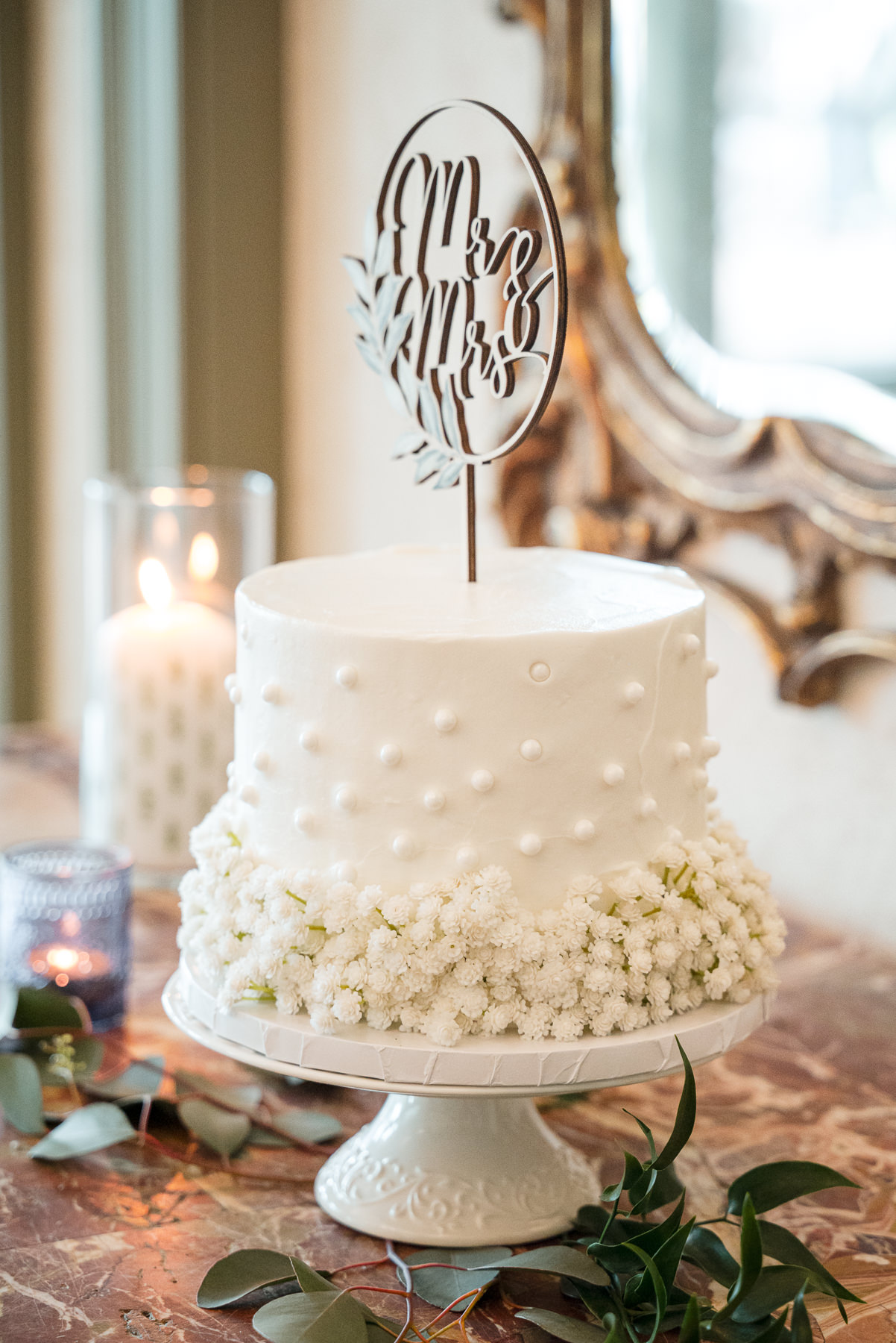 Elegant white wedding cake with pearl detailing and fresh baby’s breath flowers, topped with a “Mr & Mrs” cake topper, displayed at a spring wedding reception at Daniel Stowe Botanical Garden in Belmont, NC.