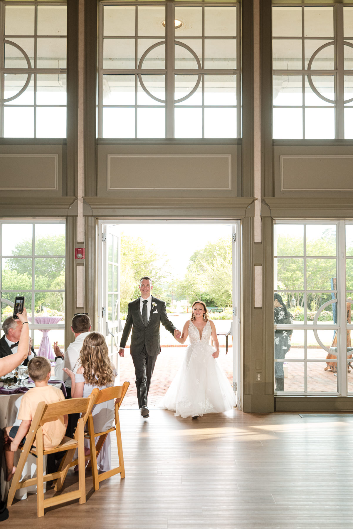 Bride and groom make their grand entrance into their wedding reception at Daniel Stowe Botanical Garden in Belmont, NC, greeted by guests inside the elegant garden ballroom.