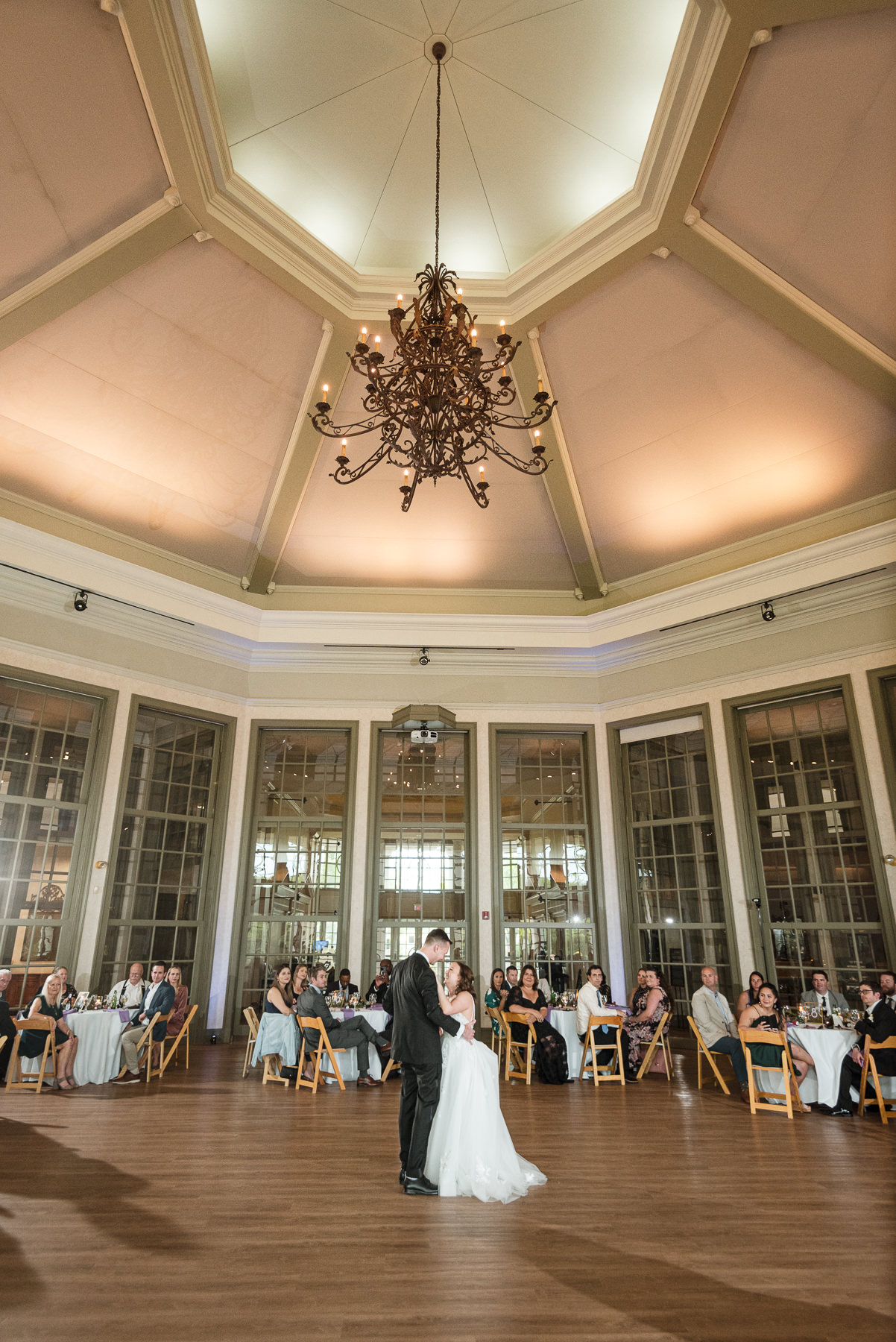Bride and groom share their first dance beneath a grand chandelier inside the ballroom at Daniel Stowe Botanical Garden in Belmont, NC, surrounded by family and friends.