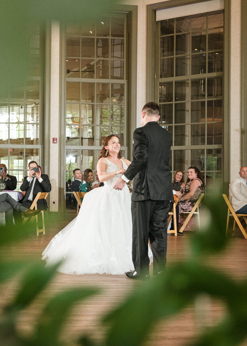 Bride and groom sharing their first dance at a spring wedding reception at Daniel Stowe Botanical Gardens in Belmont, NC, photographed by DeLong Photography, with guests watching inside the glass conservatory.