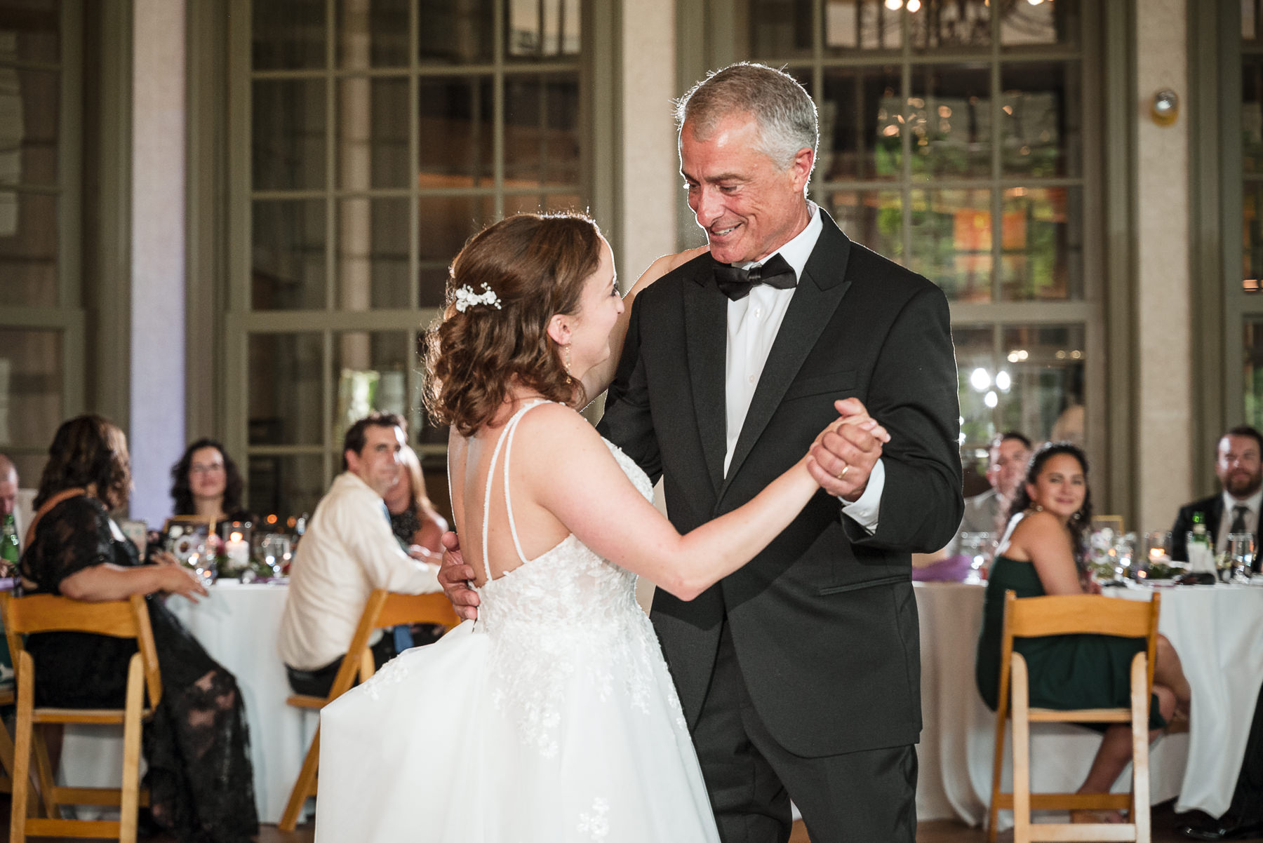 Bride sharing a father-daughter dance at a spring wedding reception at Daniel Stowe Botanical Gardens in Belmont, NC with guests watching inside the glass conservatory.
