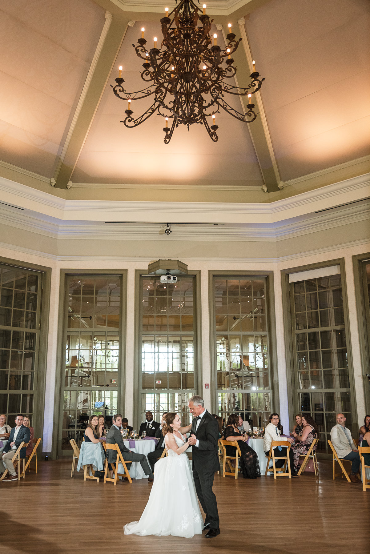 Wide view of a father-daughter dance at a spring wedding reception at Daniel Stowe Botanical Gardens in Belmont, NC, beneath a grand chandelier in the glass conservatory.