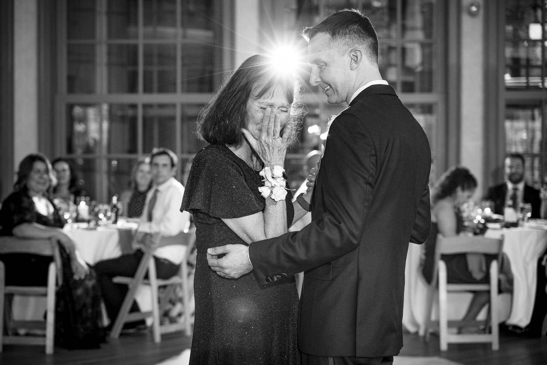 Black-and-white photo of a groom sharing an emotional mother-son dance during a spring wedding reception at Daniel Stowe Botanical Garden in Belmont, NC. Captured by DeLong Photography, this candid Charlotte wedding moment features the groom embracing his tearful mother beneath ballroom lights as guests look on.