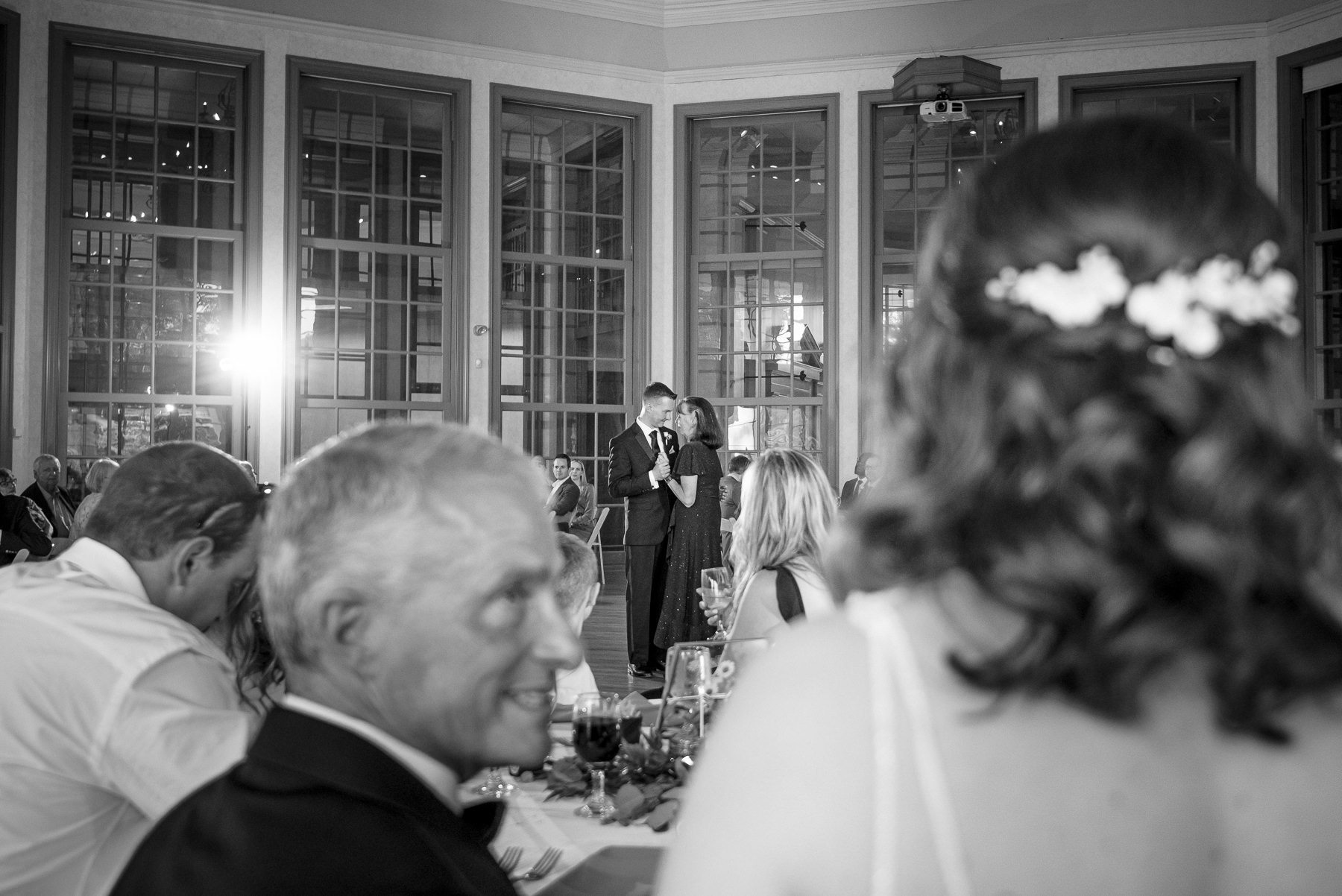 Black-and-white photo of a groom sharing a heartfelt mother-son dance during a spring wedding reception at Daniel Stowe Botanical Garden in Belmont, NC, as guests watch from their tables.