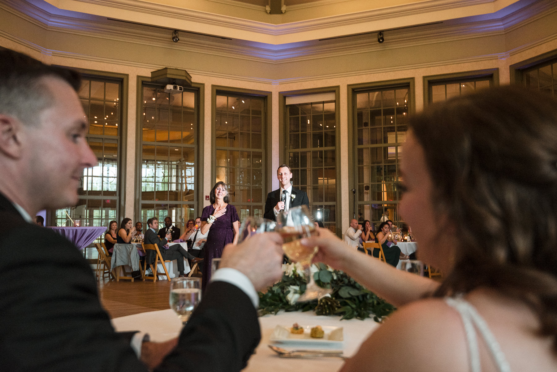 Bride and groom raise a champagne toast during their spring wedding reception at Daniel Stowe Botanical Garden in Belmont, NC, as the groom’s mother and guests look on in the elegant ballroom. Captured by DeLong Photography, this Charlotte wedding photo highlights heartfelt speeches, warm evening lighting, and the venue’s tall windows and classic architecture.
