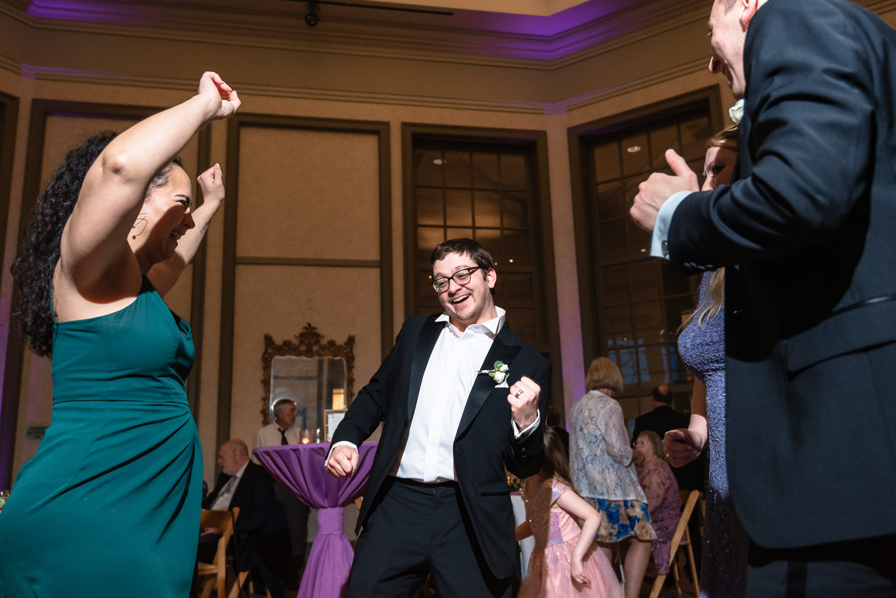 Wedding guests dancing at a spring wedding reception at Daniel Stowe Botanical Gardens in Belmont, NC, with purple uplighting inside the glass conservatory.
