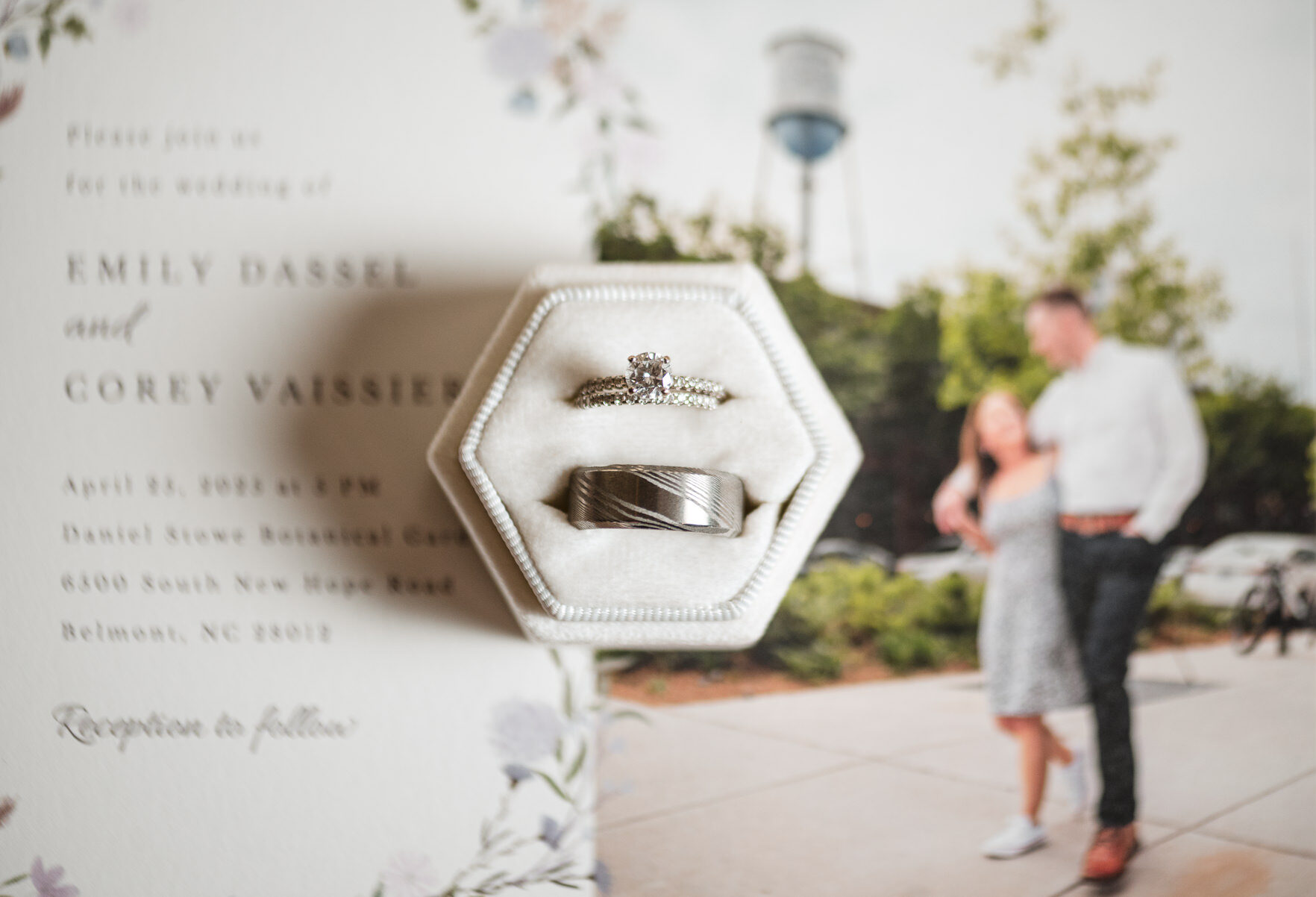 Close-up of wedding rings in a velvet box styled on an invitation suite at a spring wedding at Daniel Stowe Botanical Garden in Belmont, NC, with the couple softly blurred in the background.