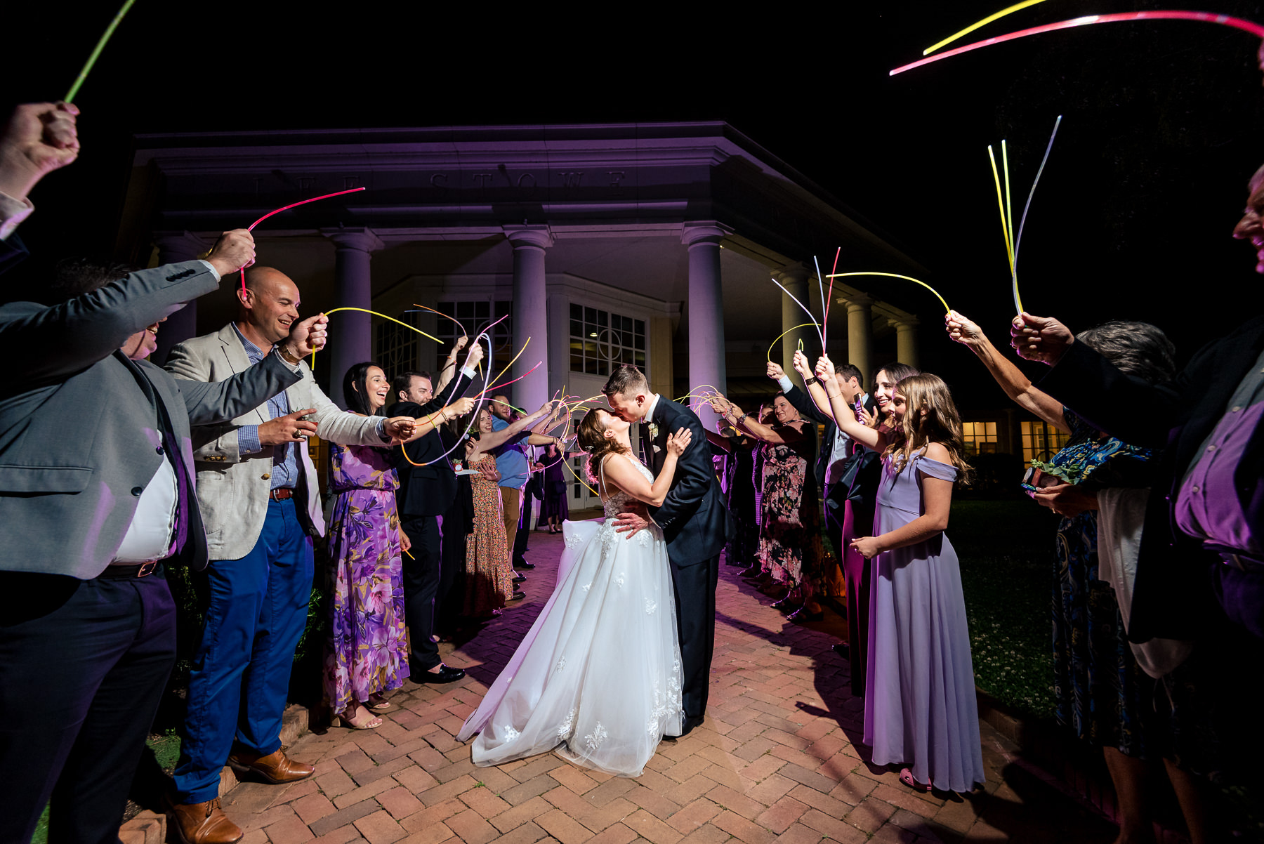 Bride and groom share a kiss during a colorful glow stick send-off outside Daniel Stowe Botanical Garden in Belmont, NC at their spring wedding reception. Captured by DeLong Photography, this vibrant Charlotte wedding exit photo features guests lining the brick walkway beneath the venue’s grand columns at night.
