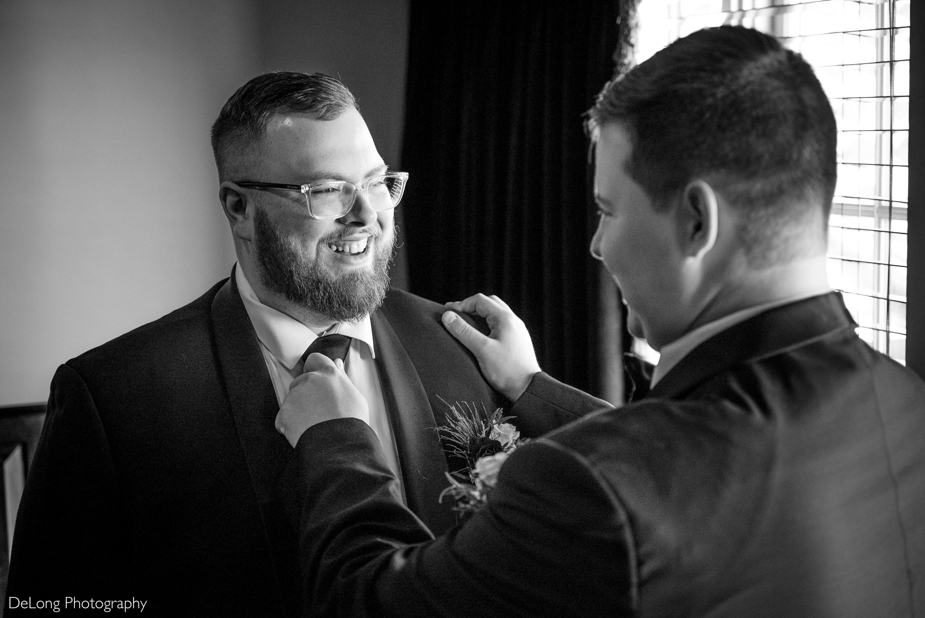 Black and white wedding getting-ready moment at Childress Vineyards in Lexington, North Carolina, featuring the groom smiling as his best man adjusts his tie before the ceremony.
