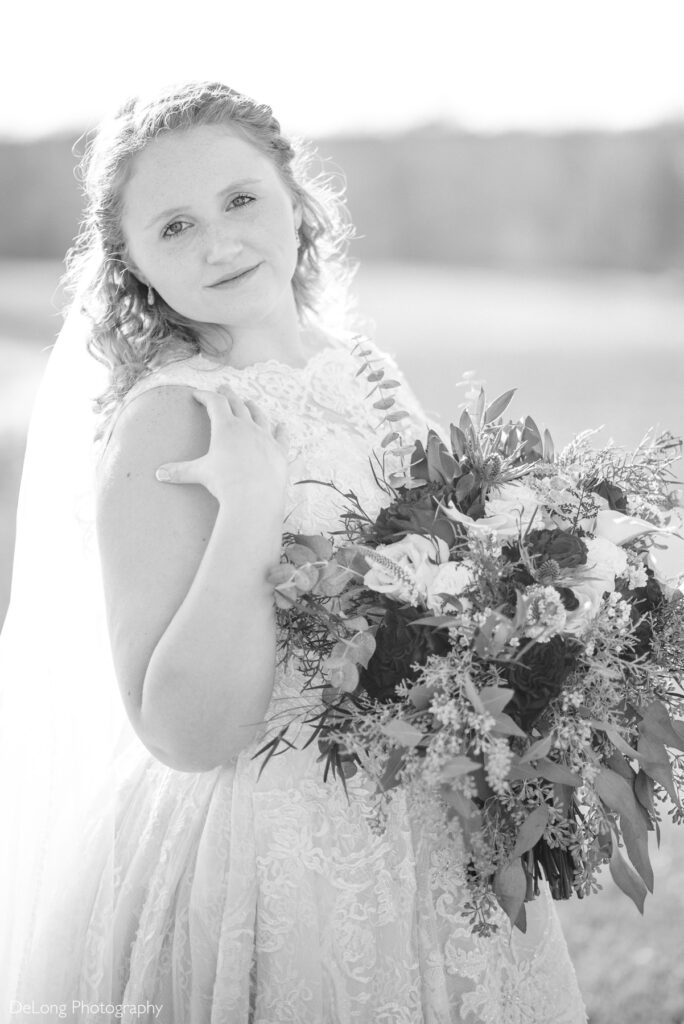 Black and white bridal portrait at Childress Vineyards in Lexington, North Carolina, featuring a bride in a lace wedding gown holding a lush floral bouquet outdoors. Soft natural light and a vineyard backdrop create a timeless, romantic wedding portrait.