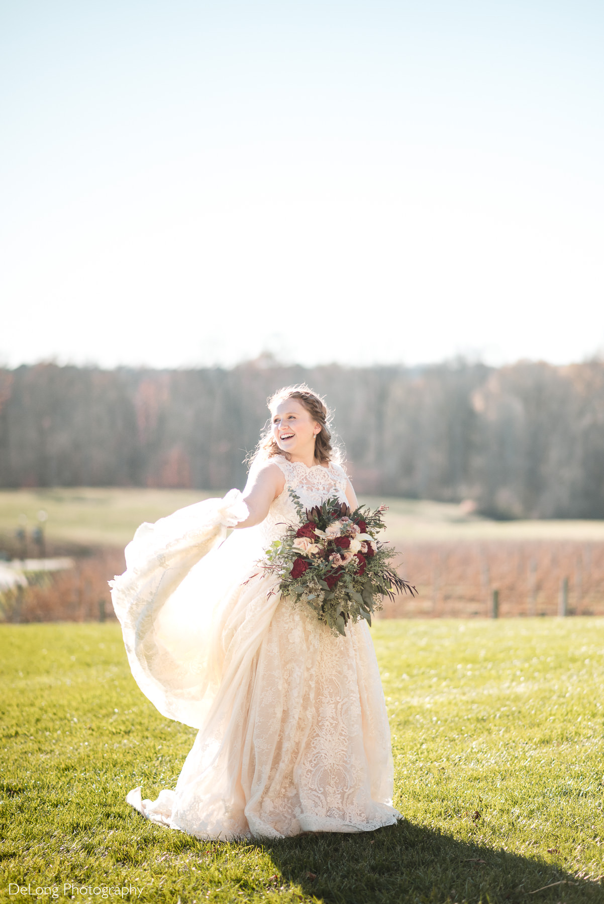Outdoor bridal portrait at Childress Vineyards in Lexington, North Carolina, featuring a joyful bride in a lace wedding gown twirling in a sunlit vineyard field while holding a rich burgundy and greenery bouquet. Rolling vineyards and soft natural light create a romantic, scenic backdrop for this Childress Vineyards wedding.