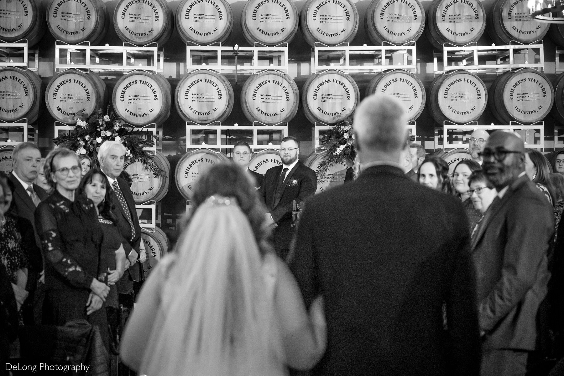 Indoor wedding ceremony at Childress Vineyards in Lexington, North Carolina, with guests gathered between wine barrels, photographed by DeLong Photography.