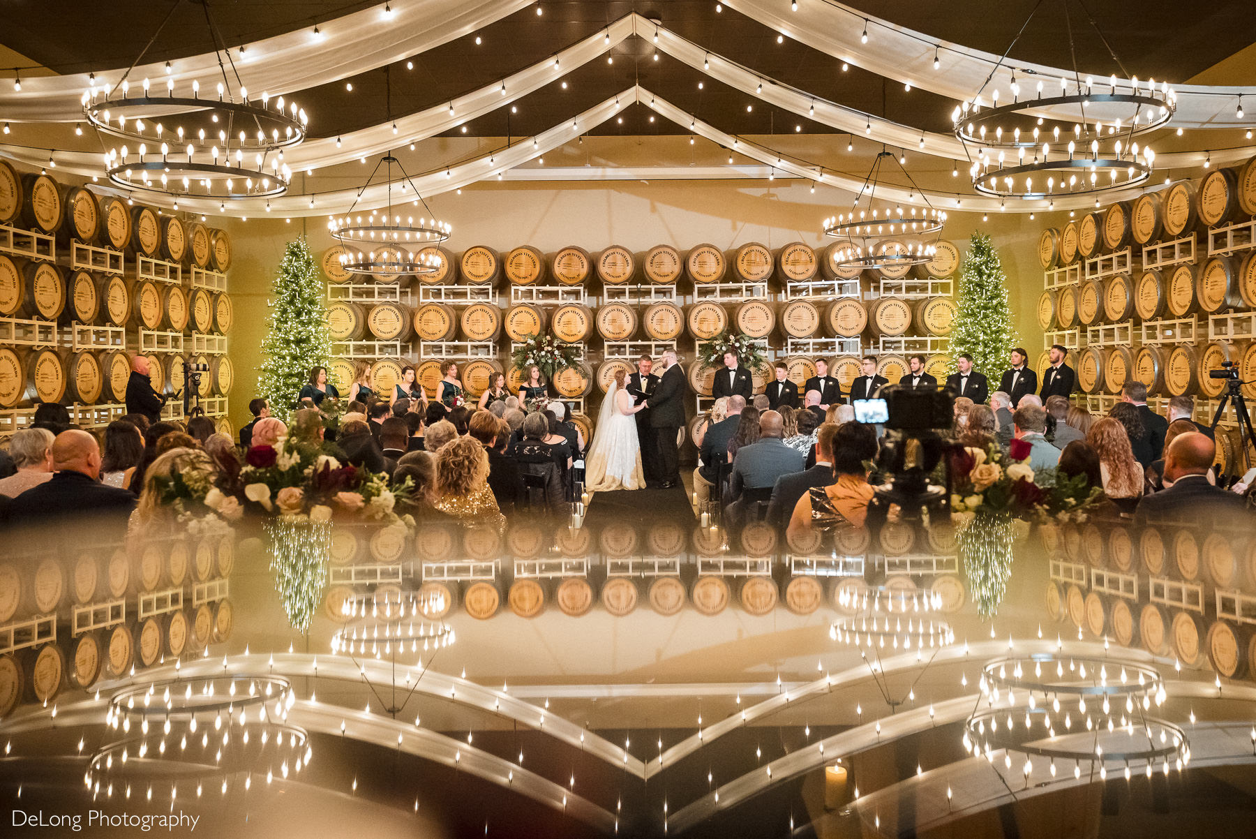 Wedding ceremony in the wine cellar at Childress Vineyards in Lexington, North Carolina, featuring chandeliers, Christmas trees, and rows of wine barrels, photographed by DeLong Photography.