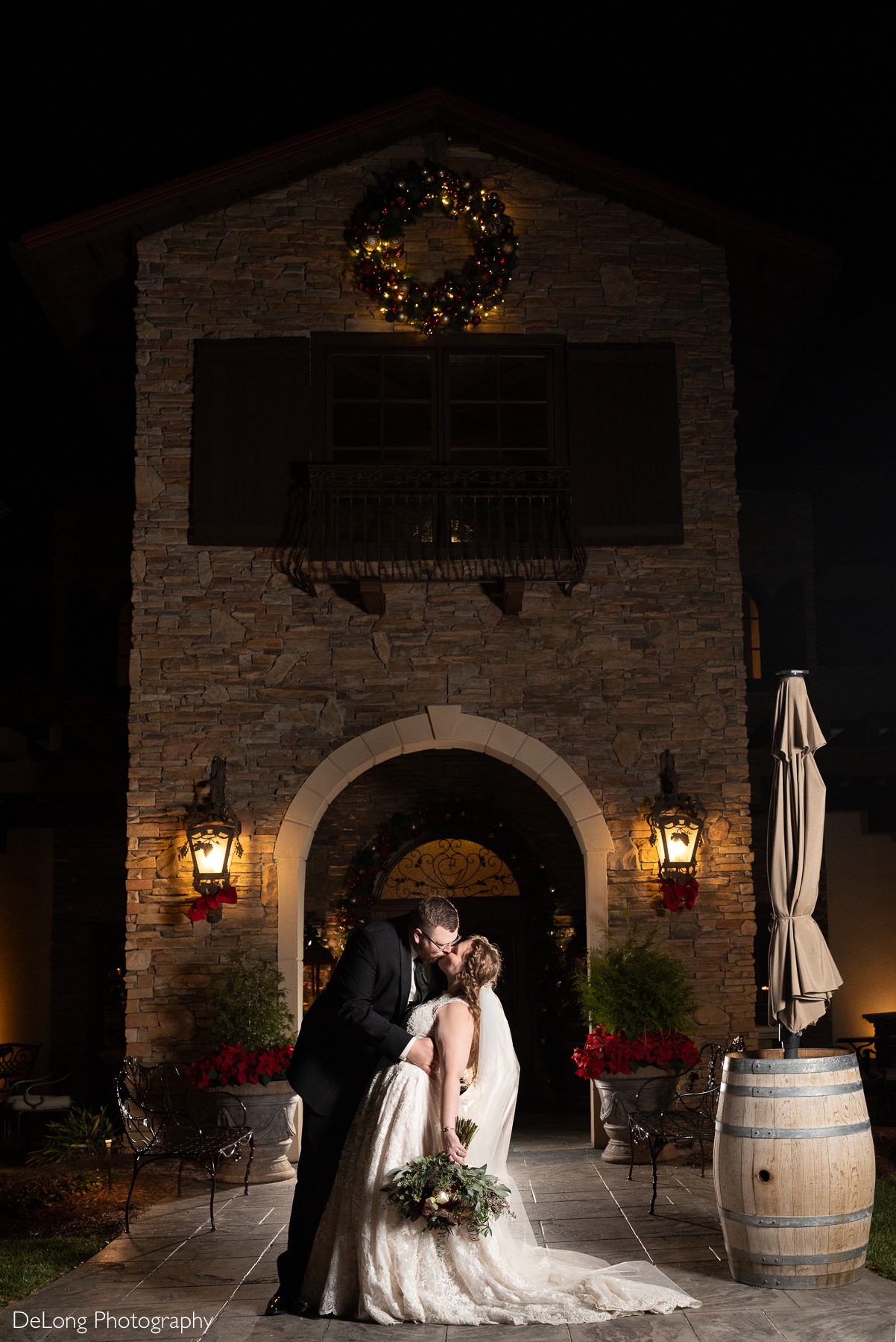 Bride and groom share a romantic night time kiss beneath a holiday wreath at Childress Vineyards in Lexington, North Carolina. Captured by DeLong Photography, this image showcases elegant winter wedding portraits with dramatic vineyard architecture and festive lighting.