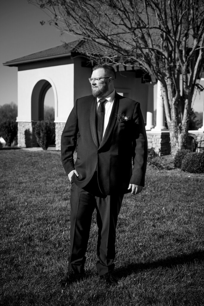 Black and white portrait of the groom standing outdoors at Childress Vineyards in Lexington, North Carolina, wearing a classic black suit and boutonniere on his wedding day, photographed by DeLong Photography.