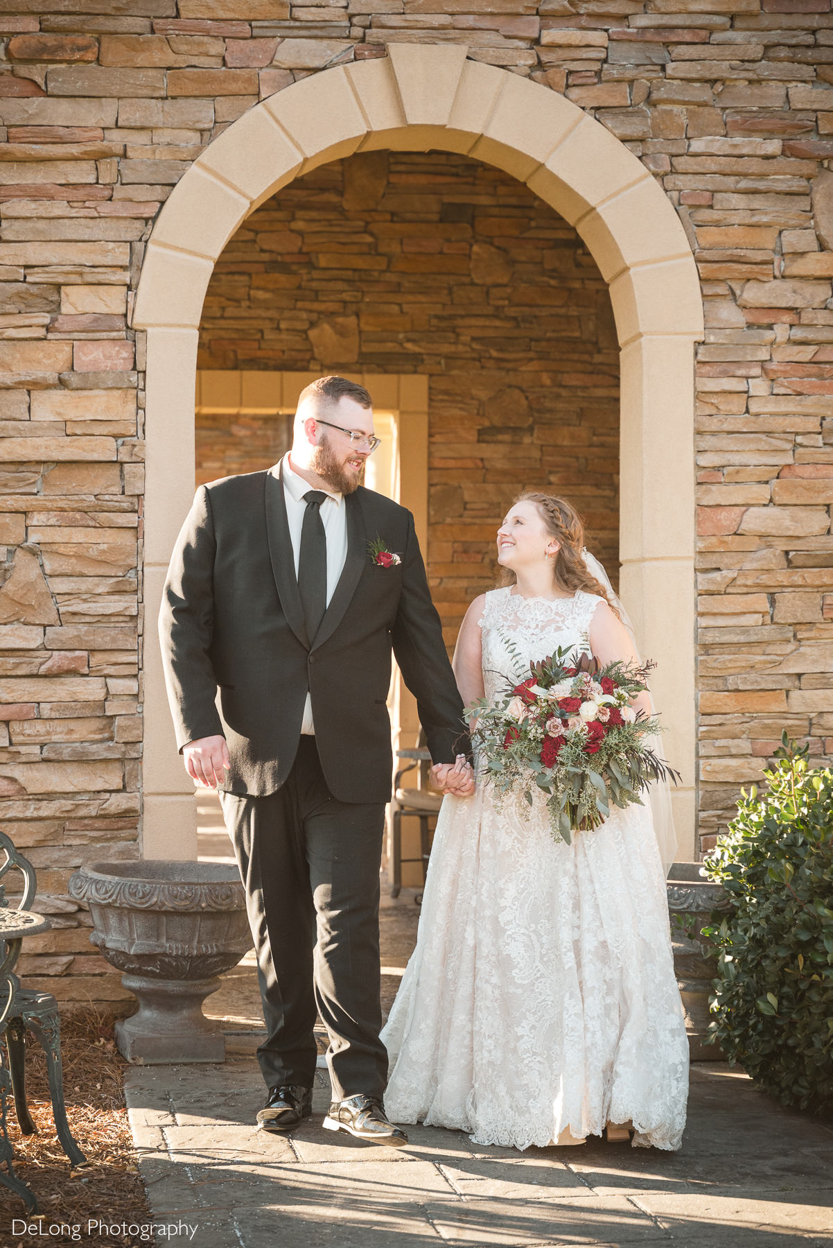 Bride and groom walking hand in hand beneath a stone archway at Childress Vineyards in Lexington, North Carolina, sharing a joyful moment during their wedding portraits.