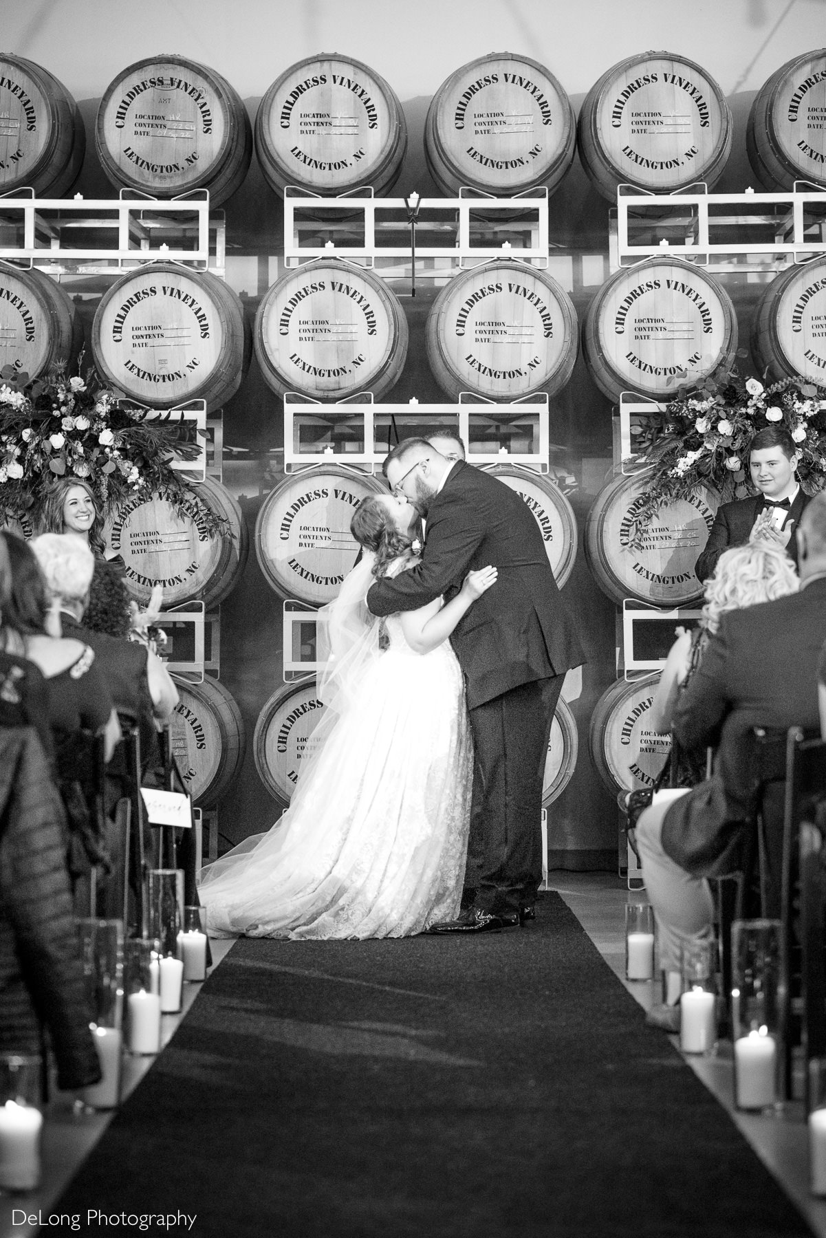 Bride and groom sharing their first kiss during an indoor winery wedding ceremony at Childress Vineyards in Lexington, North Carolina, framed by wine barrels and candlelit aisle.