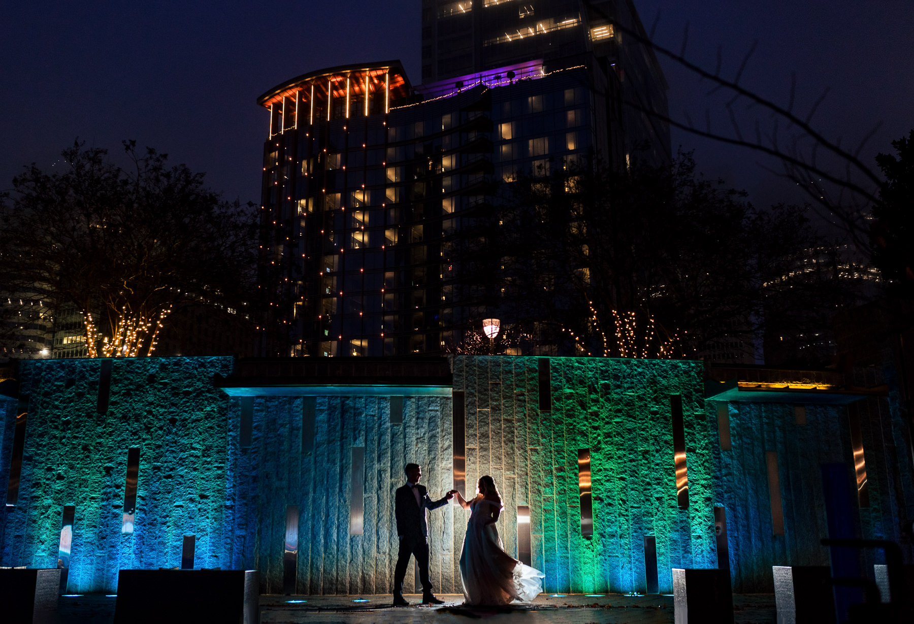 Nighttime silhouette of bride and groom dancing in front of the water wall in Romare Bearden Park that is lit blue and green, The twinkling lights of the city skyline and deep blue sky is beyond this.