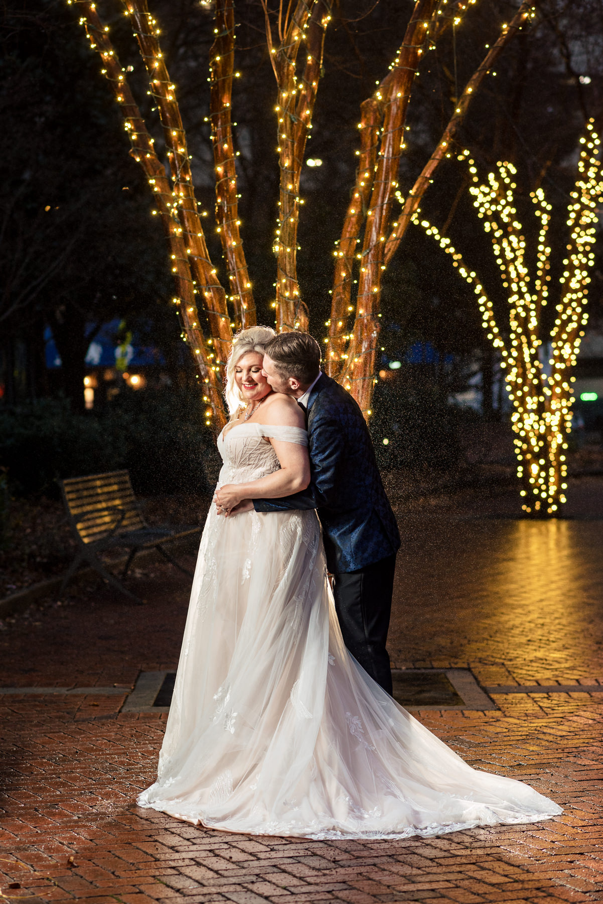 Bride and groom embrace during a rainy evening elopement portrait session at Romare Bearden Park in Uptown Charlotte, NC, surrounded by glowing string lights and city ambiance.