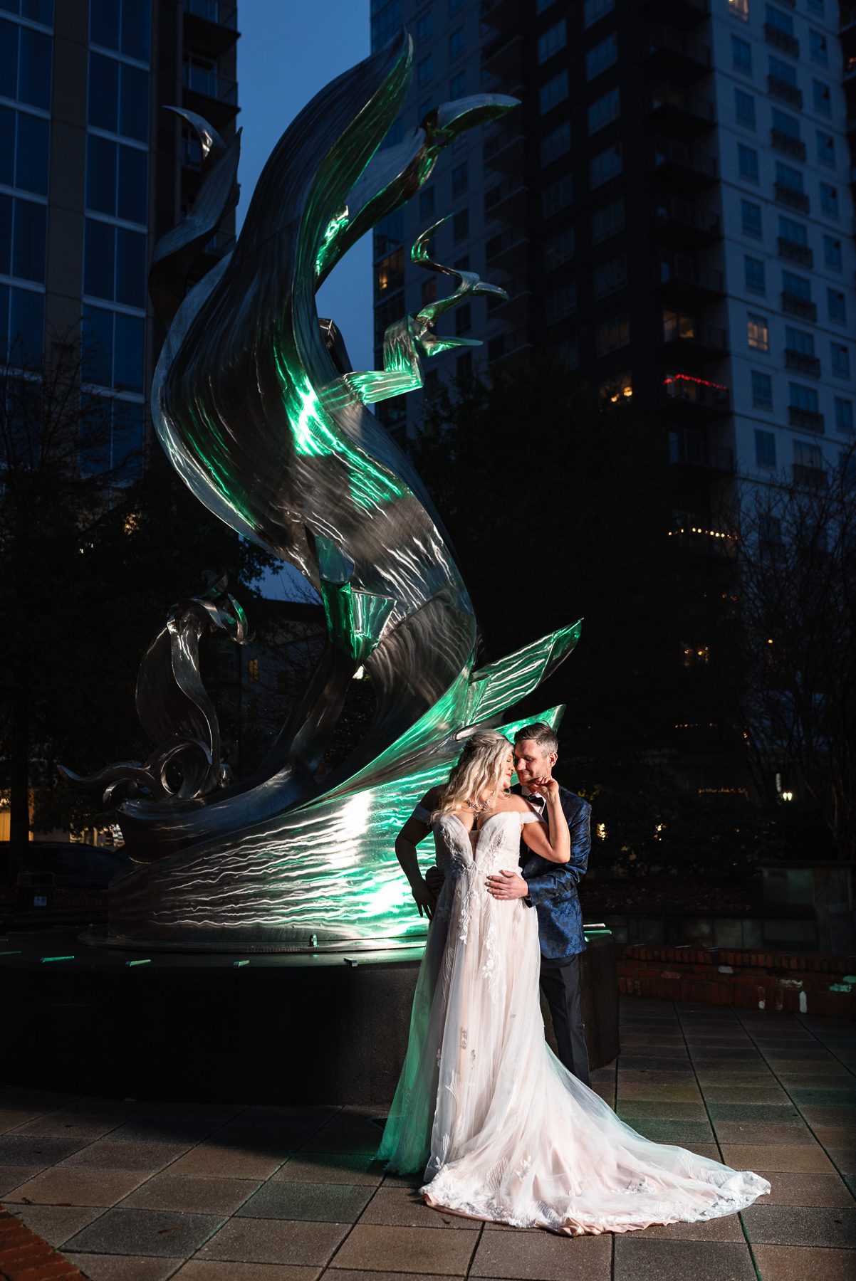 Bride and groom pose for dramatic evening elopement portraits at Romare Bearden Park in Uptown Charlotte, NC, standing beneath a modern illuminated sculpture with the city skyline behind them.