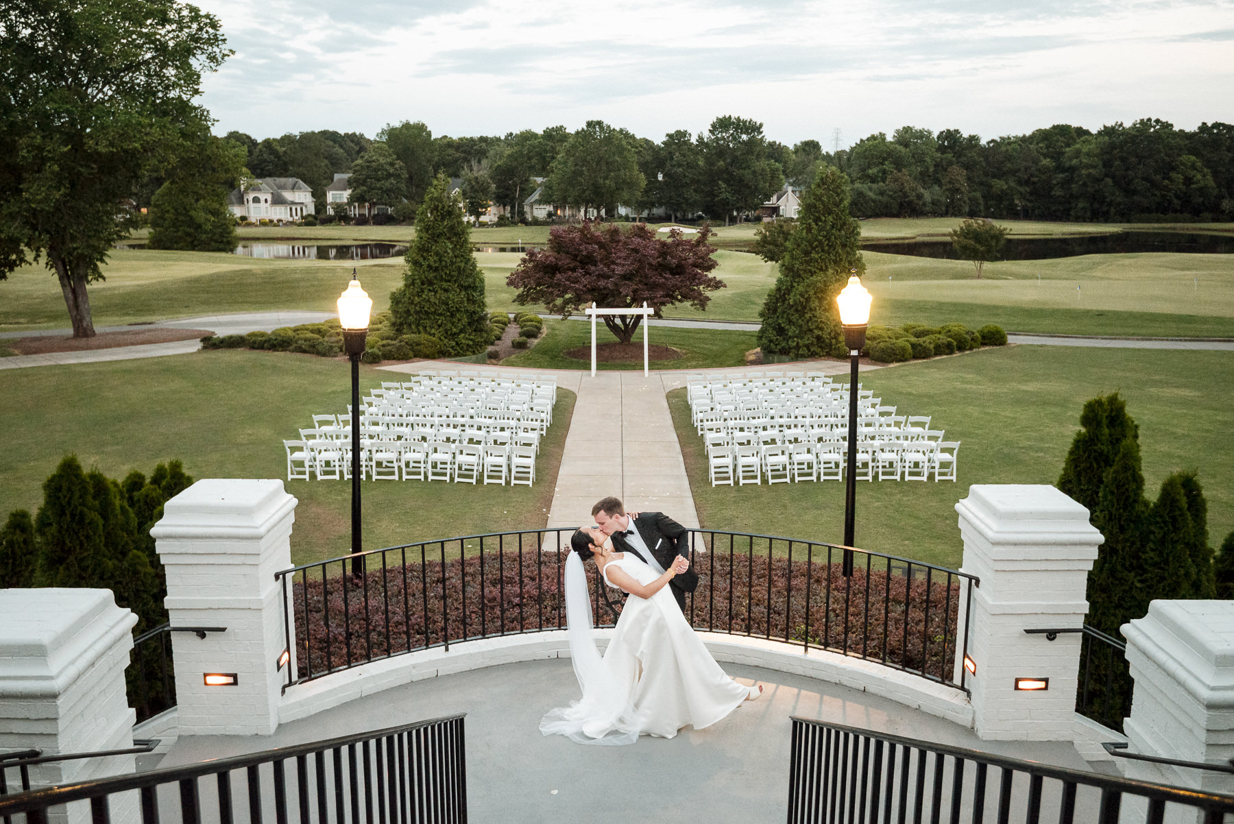 Bride and groom share a romantic kiss on the terrace overlooking the ceremony setup at Providence Country Club in Charlotte, North Carolina. Elegant outdoor wedding photography by DeLong Photography highlights the scenic golf course, symmetrical aisle, and classic country club architecture.