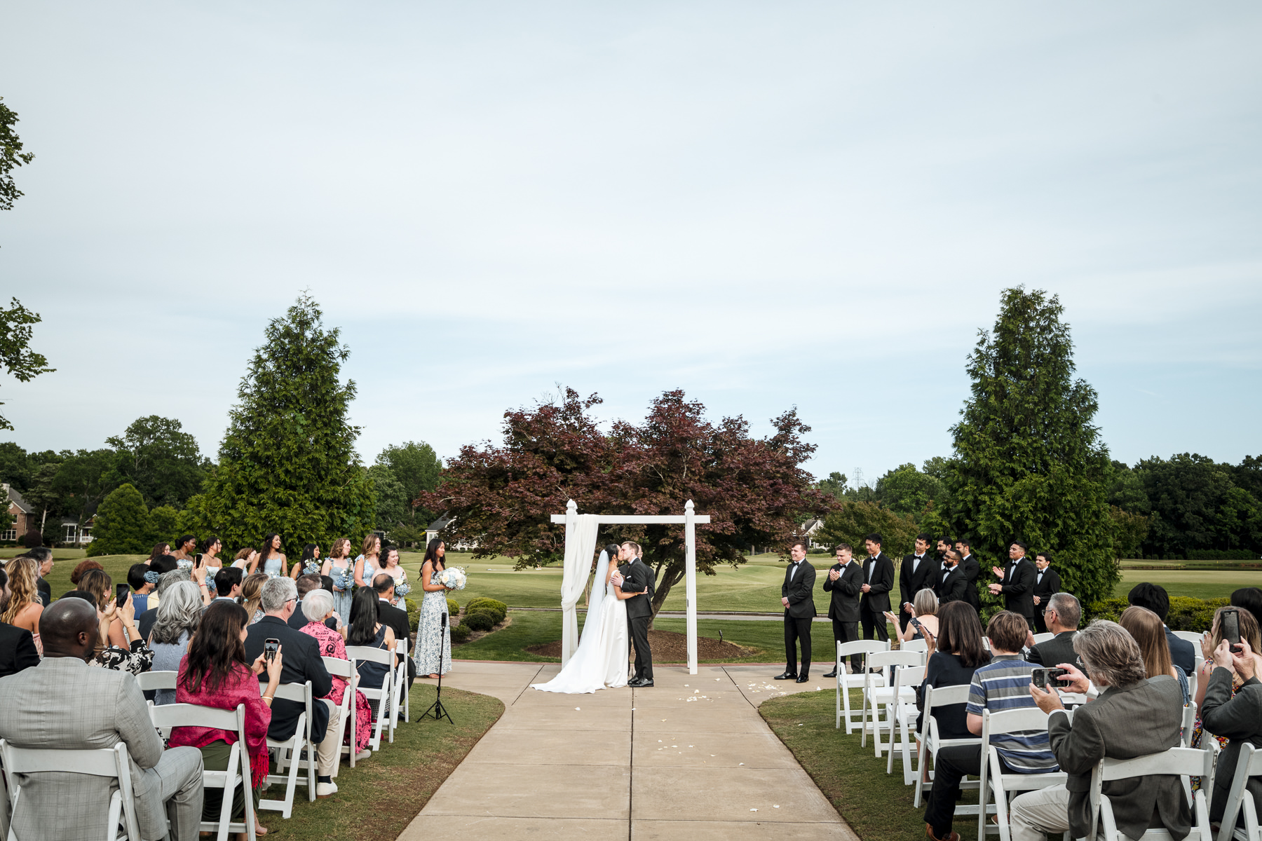 Bride and groom share their first kiss during an outdoor wedding ceremony at Providence Country Club in Charlotte, NC, photographed by DeLong Photography. This elegant golf course wedding showcases the timeless, candid moments couples look for when searching for a Charlotte wedding photographer.
