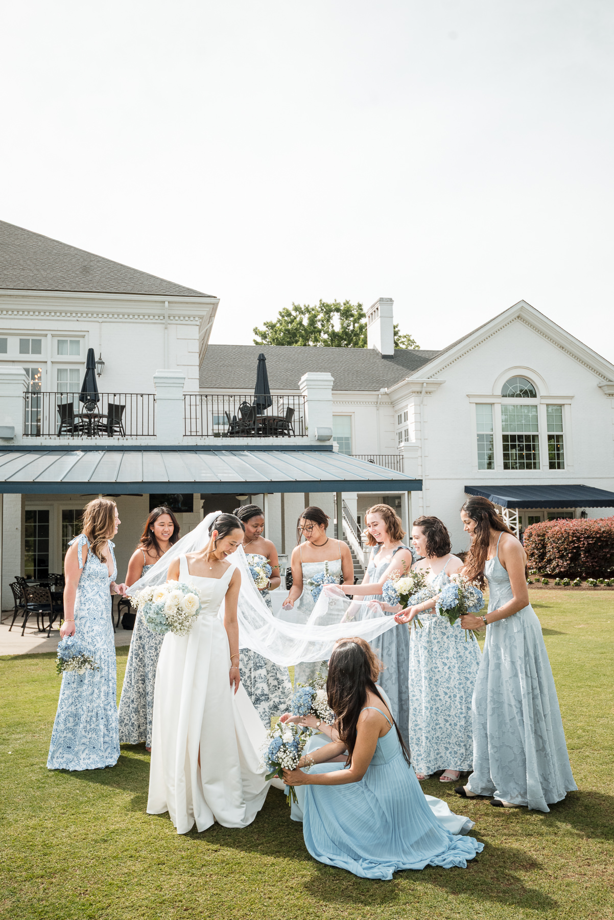 Bridesmaids portrait at Providence Country Club in Charlotte, NC, featuring the bride and her wedding party in blue dresses gathered around her veil on the lawn, captured by DeLong Photography.
