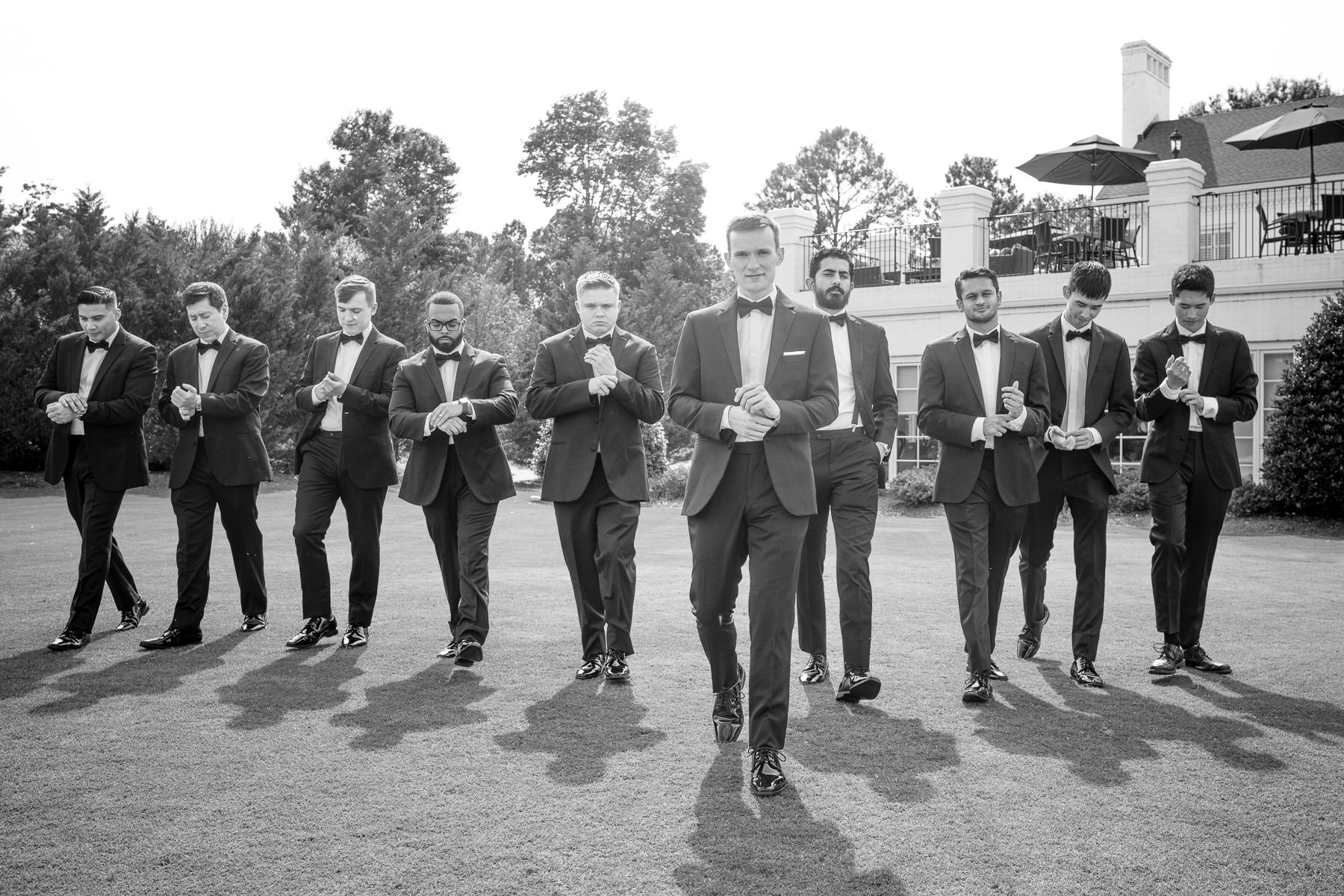 Groomsmen in black tuxedos walking together across the lawn at Providence Country Club in Charlotte, North Carolina during a wedding day, captured in a classic black and white portrait by DeLong Photography.
