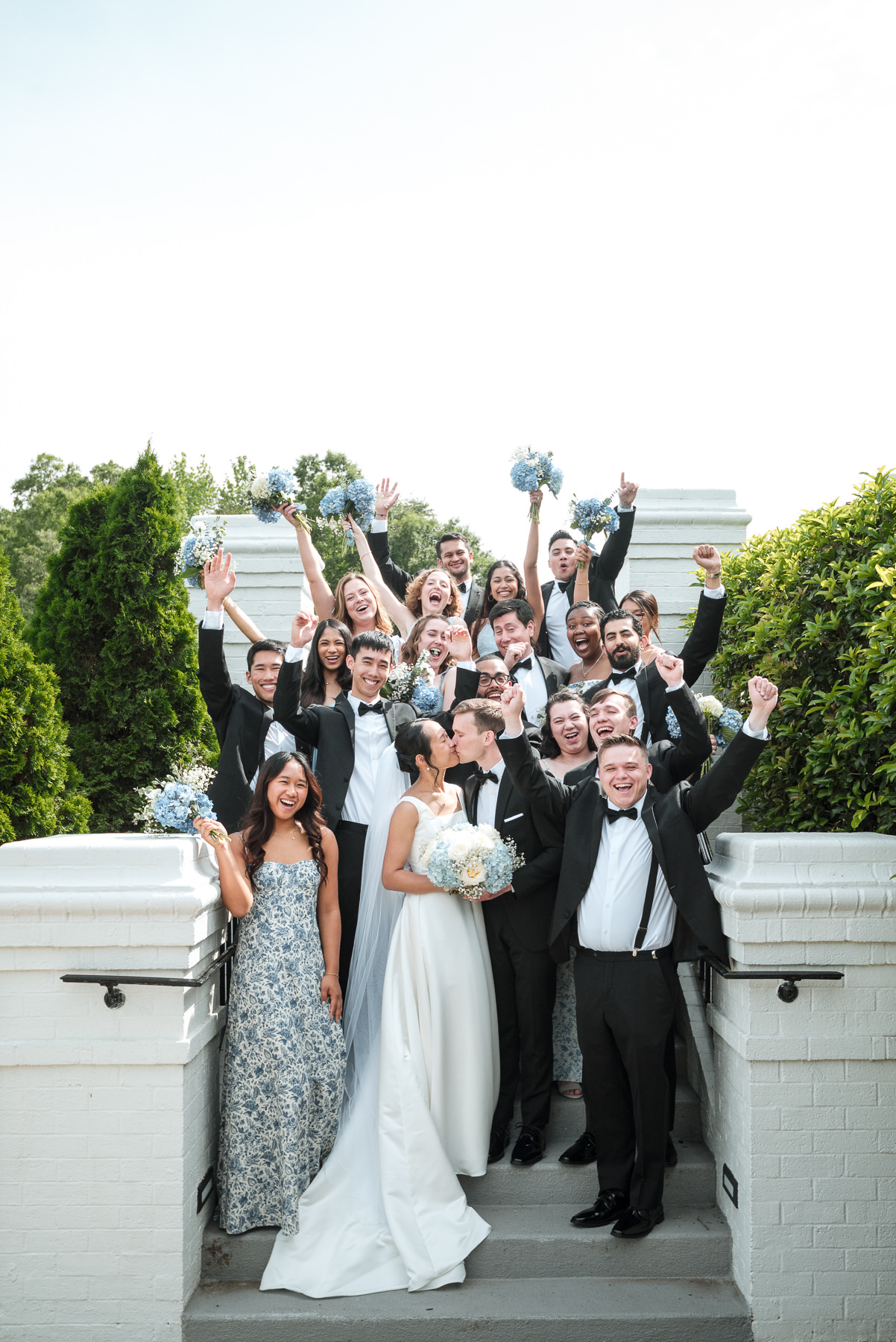 Wedding party celebration at Providence Country Club in Charlotte, NC. The bride and groom share a kiss while surrounded by cheering bridesmaids and groomsmen holding blue hydrangea bouquets. Captured by DeLong Photography, Charlotte wedding photographers specializing in luxury country club weddings and joyful candid moments.