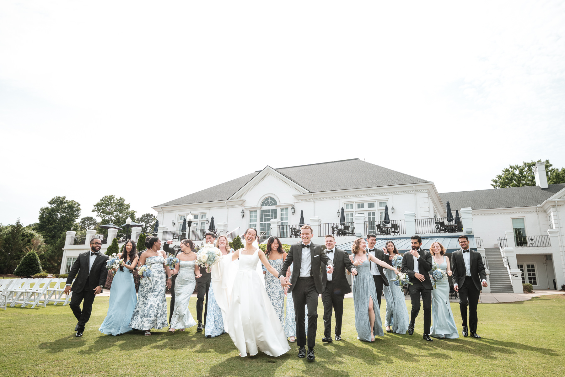 Wedding party walking joyfully across the lawn at Providence Country Club in Charlotte, NC, with the bride and groom leading bridesmaids and groomsmen in coordinated blue and black attire in front of the clubhouse. Photographed by DeLong Photography, Charlotte wedding photographers.