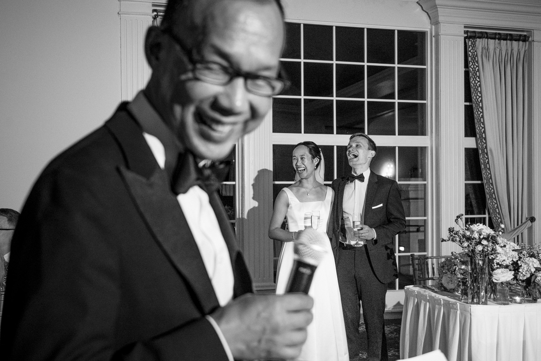 Father of the bride gives an emotional wedding toast as the bride and groom laugh together during their reception at Providence Country Club, photographed by DeLong Photography in Charlotte, NC. This candid wedding reception moment showcases the storytelling style couples look for when searching for Charlotte wedding photographers.