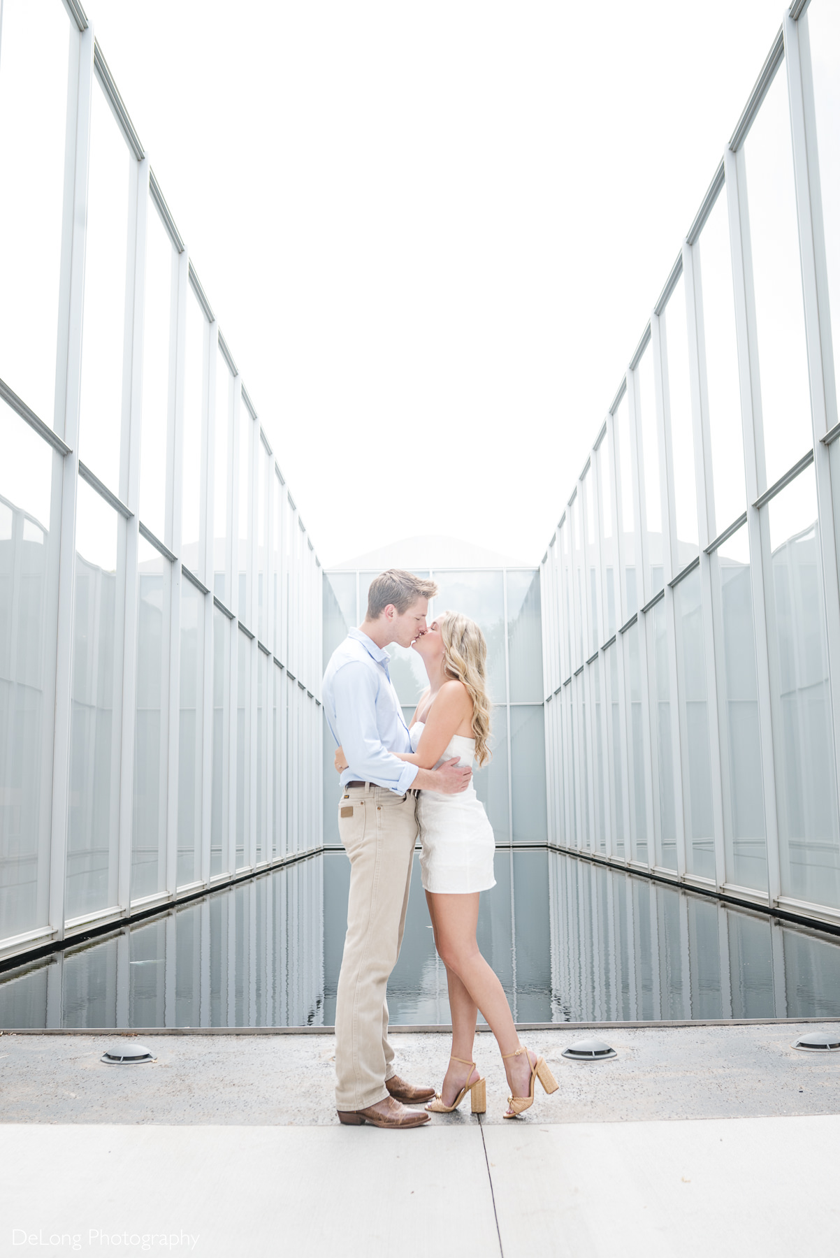 NC Art Museum engagement session in Raleigh, NC, featuring a couple kissing between the glass walls of the museum’s modern architecture, captured by DeLong Photography.