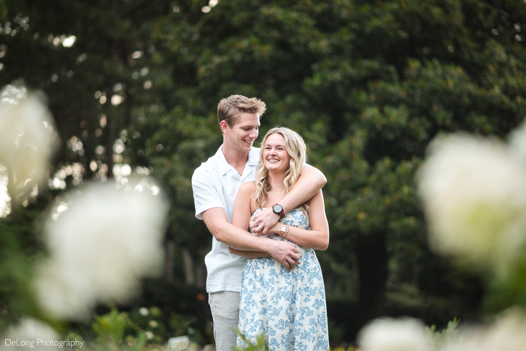 Portrait of an engaged couple during a WRAL Azalea Garden engagement session in Raleigh, NC, with the bride-to-be smiling as her fiancé giggles with his arms around her. The couple is framed by flowers in the foreground. Captured by DeLong Photography during a timeless and romantic engagement session.