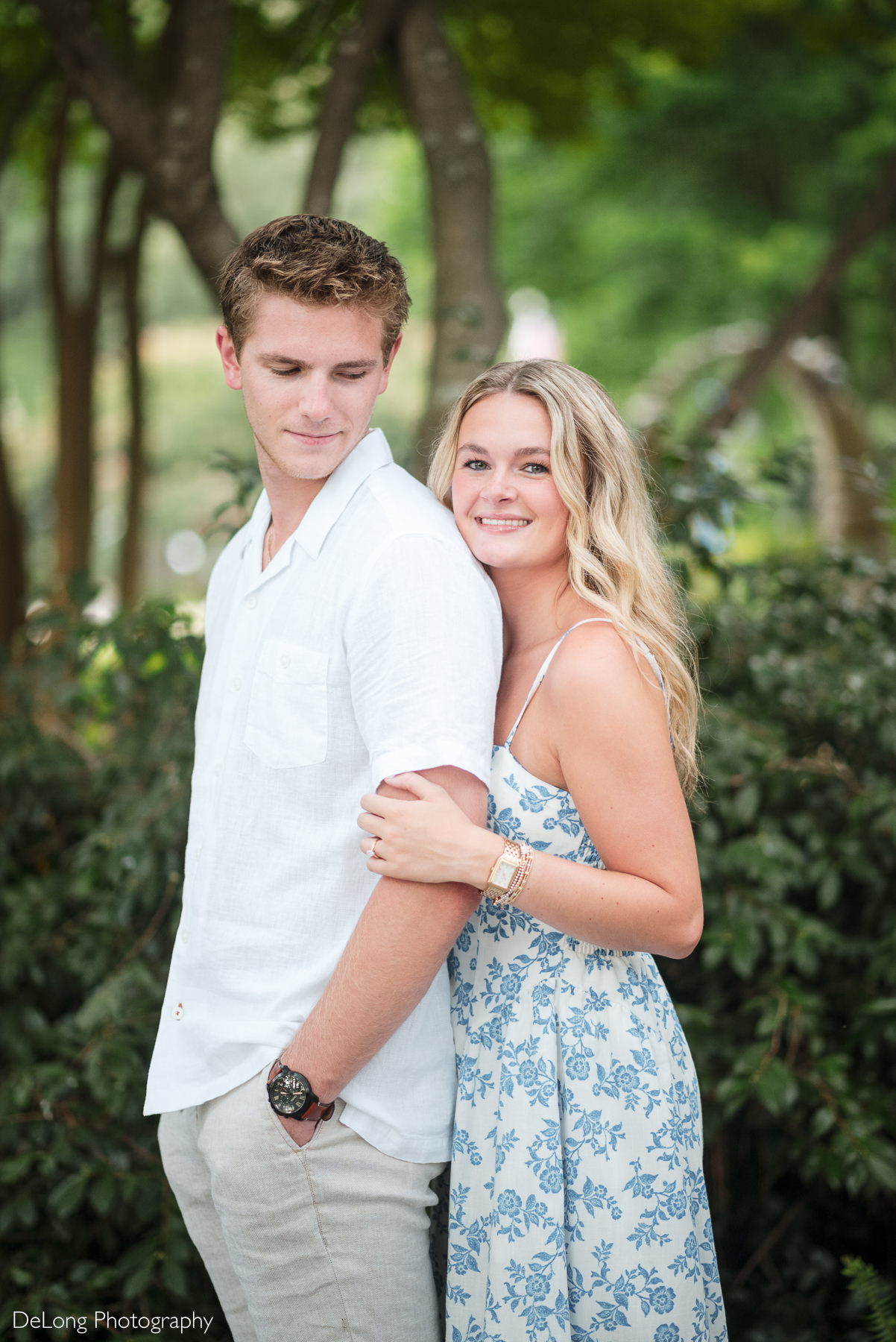 Portrait of an engaged couple during a WRAL Azalea Garden engagement session in Raleigh, NC, with the bride-to-be embracing her fiancé from behind and smiling at the camera. Captured by DeLong Photography during a timeless and romantic engagement session.