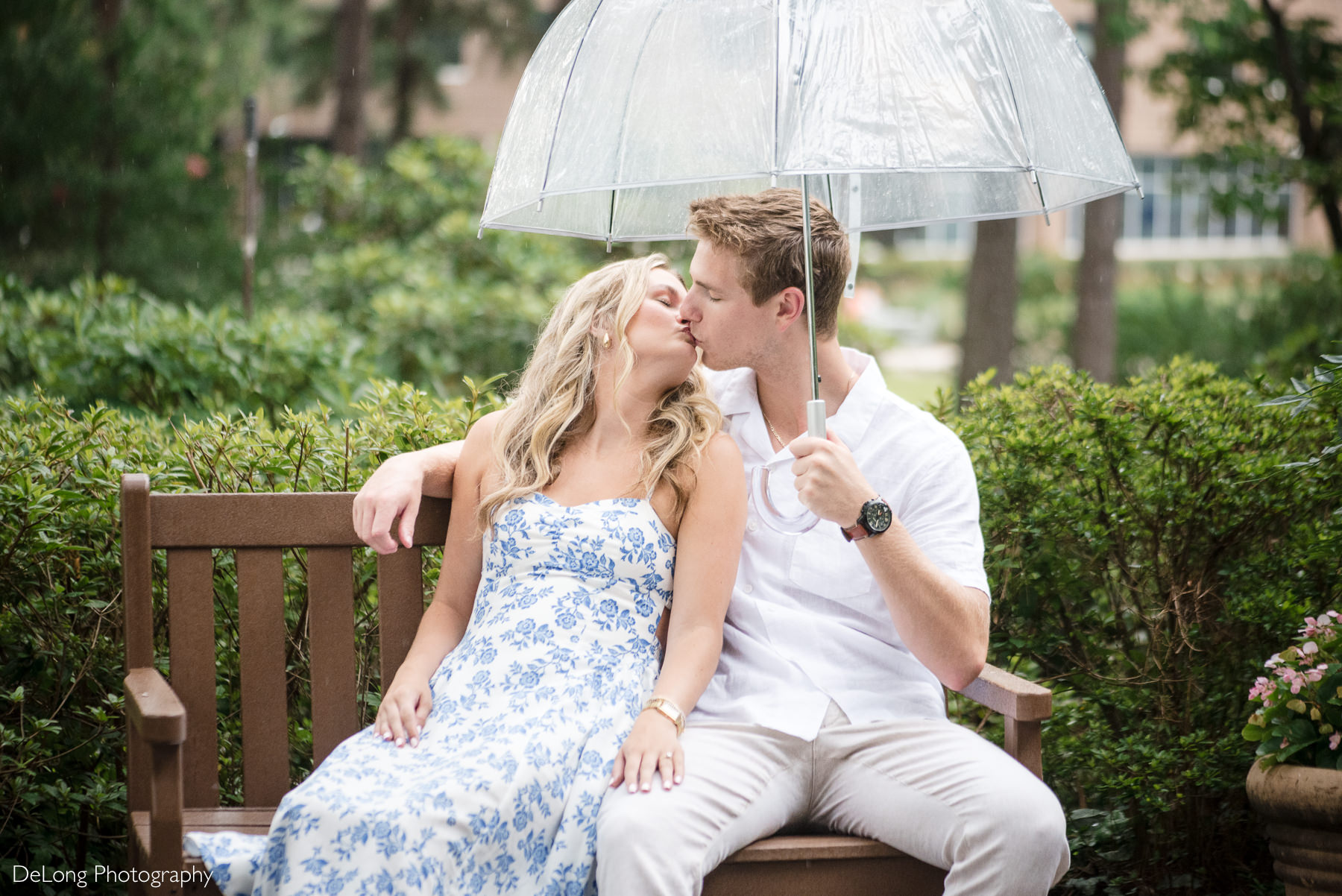 Portrait of an engaged couple during a WRAL Azalea Garden engagement session in Raleigh, NC, with the couple sharing a kiss sitting on a bench, under a clear umbrella held by the gentleman. Captured by DeLong Photography a Raleigh engagement photographer.