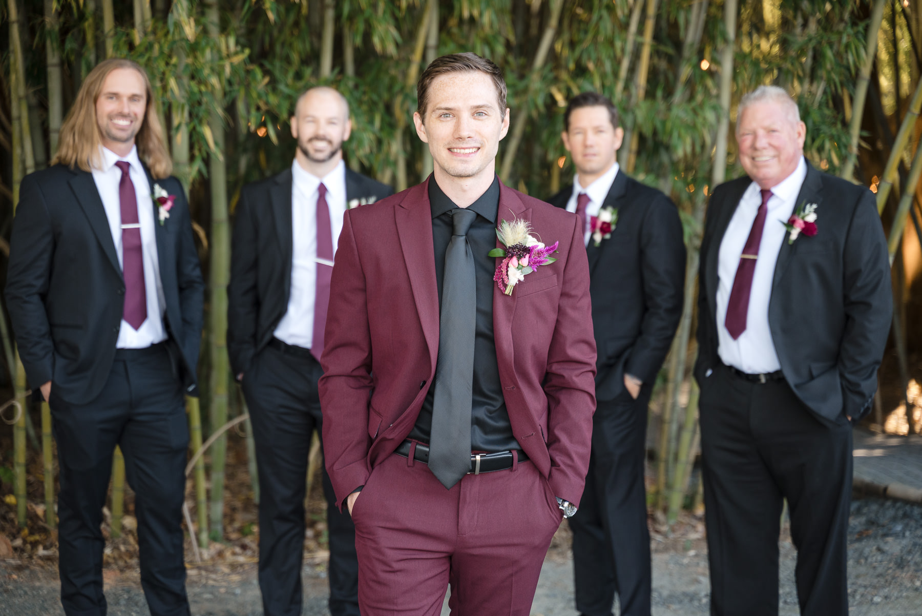 Groom in a burgundy suit with his groomsmen before the ceremony at Camelot Meadows in Charlotte, NC. Fall wedding party portrait in the venue's bamboo forest captured by DeLong Photography, a Charlotte wedding photographer.