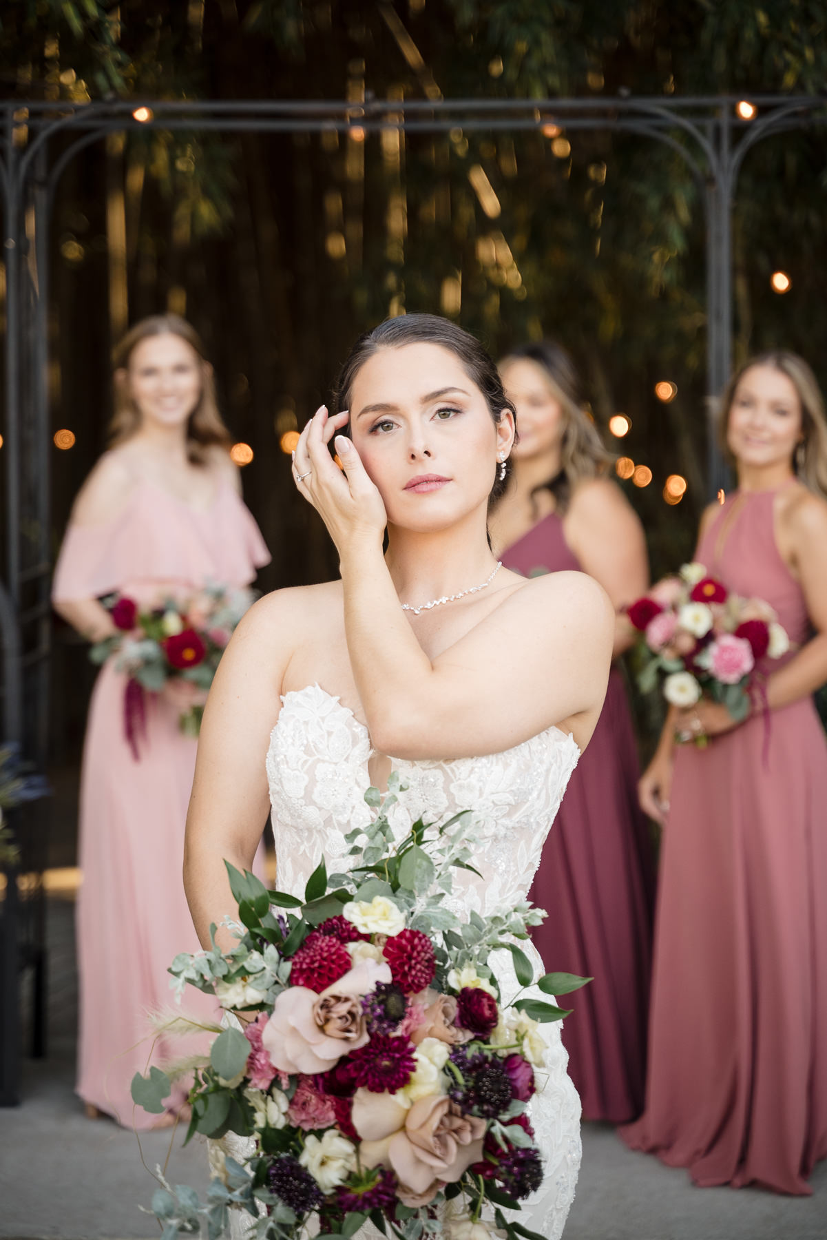 Bride holding a burgundy and blush bridal bouquet with bridesmaids standing behind her in the bamboo forest at Camelot Meadows in Charlotte, NC. Elegant fall wedding party portrait captured by DeLong Photography, a Charlotte wedding photographer.