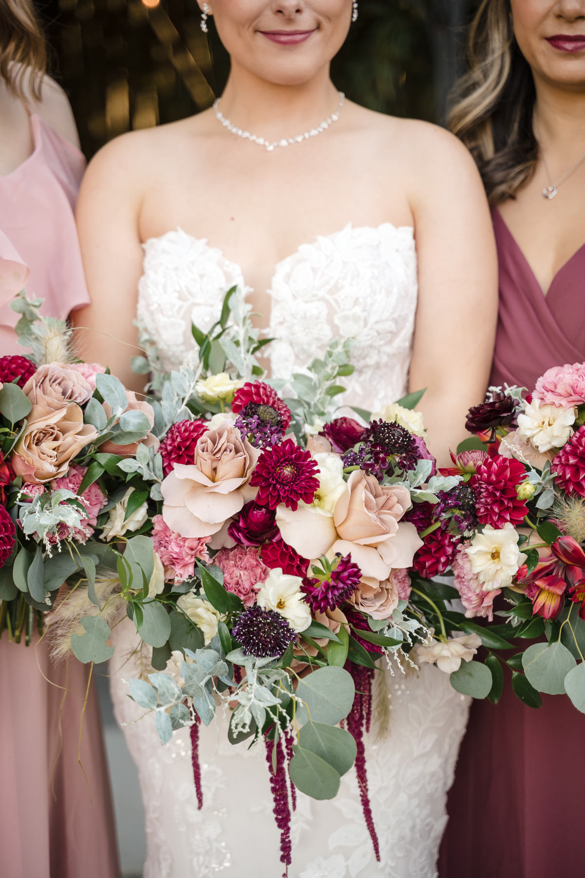 Bride and bridesmaids hold lush burgundy, blush, ivory, and eucalyptus bouquets during bridal party portraits at Camelot Meadows in Charlotte, NC. Elegant wedding flowers complement the bride's lace gown, captured by Charlotte wedding photographers DeLong Photography.